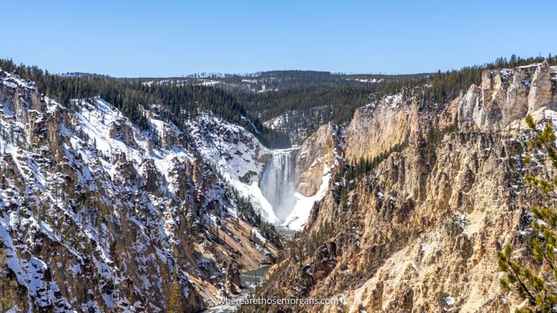 Photo of a powerful waterfall at the end of a massive V-shaped valley with light snow on the ground and a sunny sky