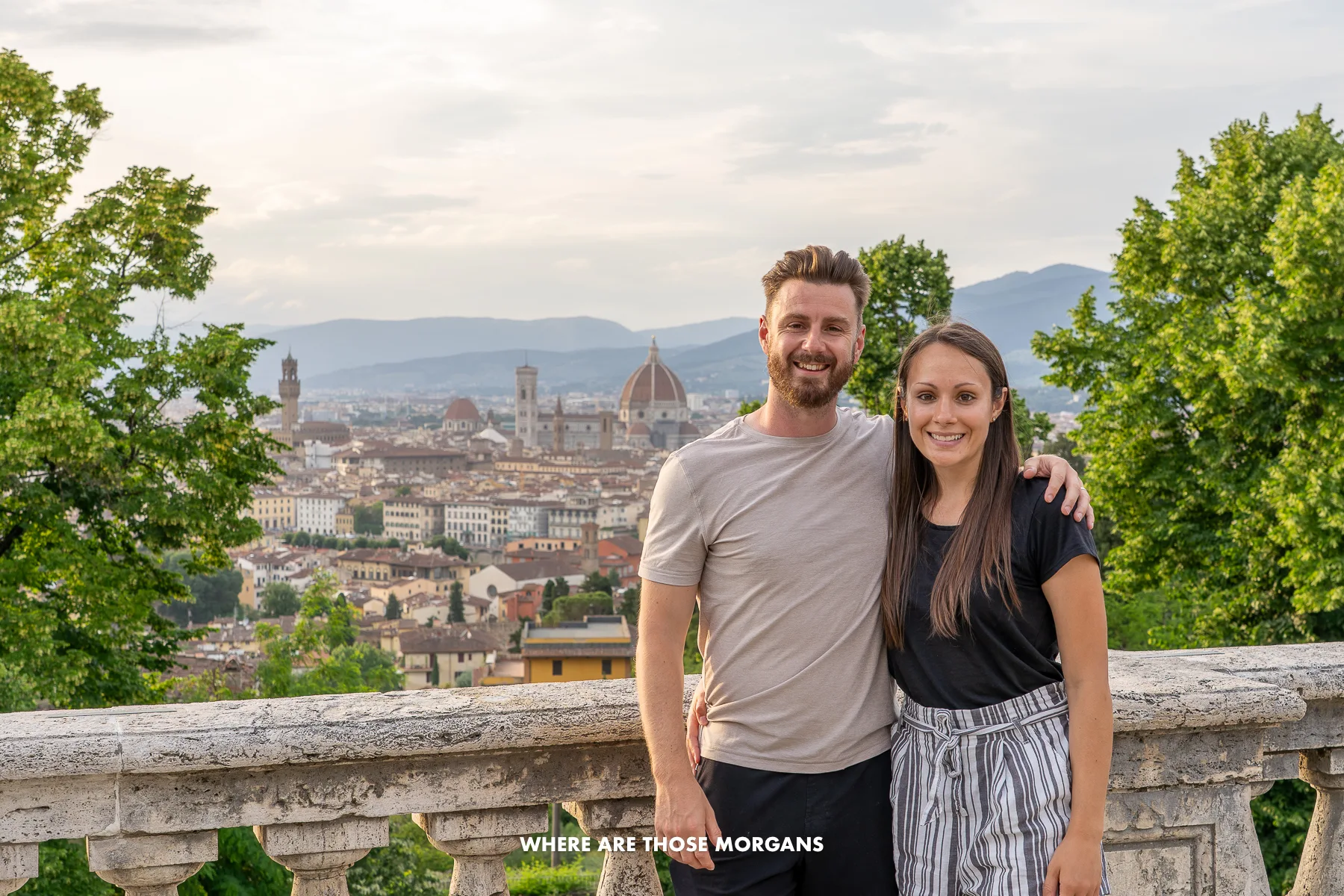 Mark and Kristen from Where Are Those Morgans having a photo taken in front of a low stone balcony with trees and views over Florence Italy behind