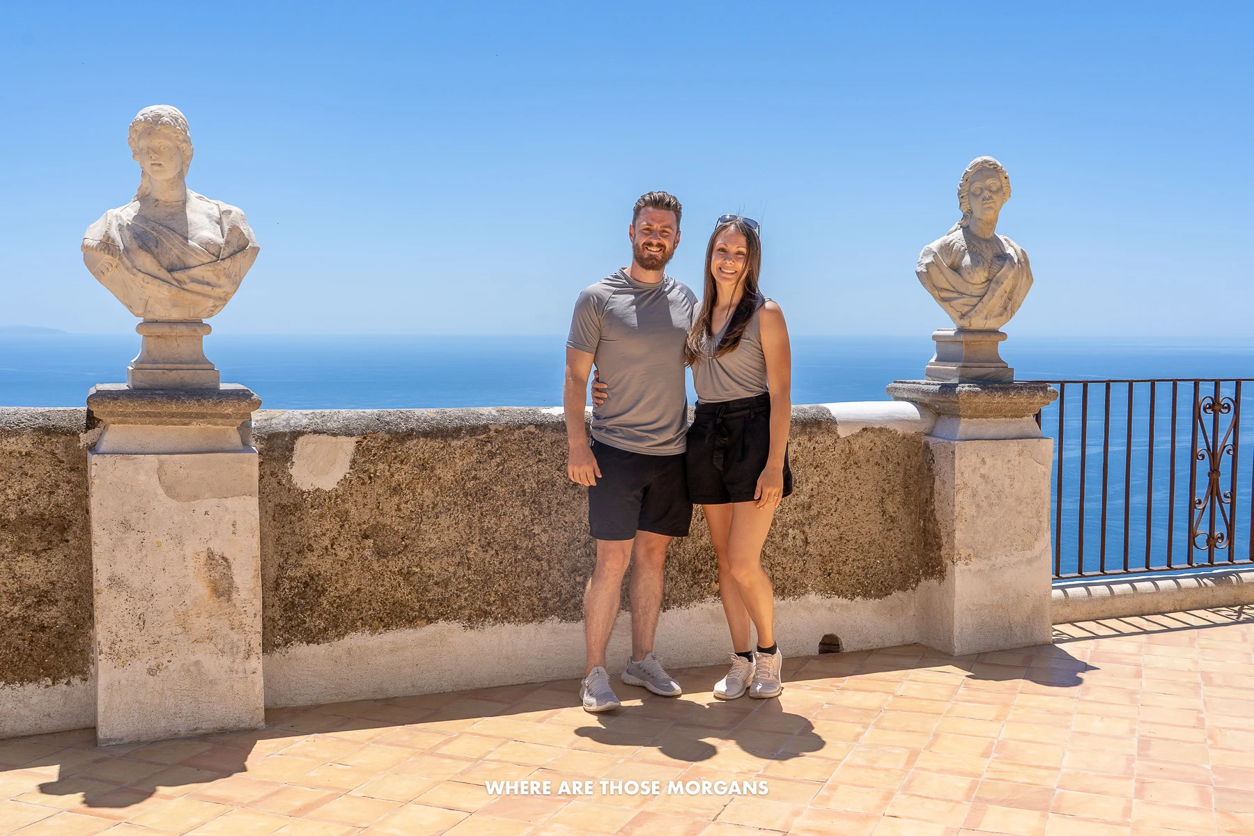 Mark and Kristen from Where Are Those Morgans in front of a low stone wall with busts and views over the ocean behind on a sunny day in Italy