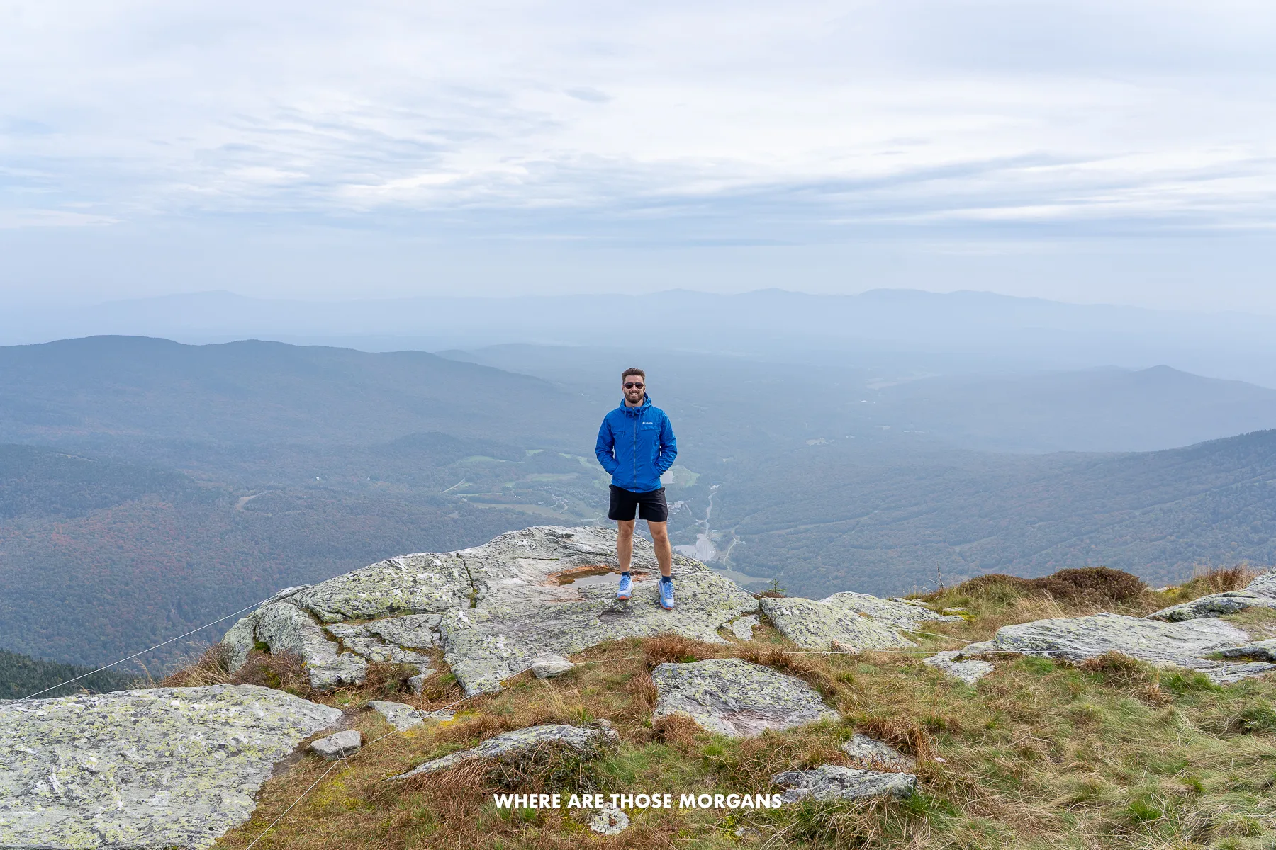 Photo of Mark Morgan from Where Are Those Morgans standing on rocks at the summit of Mt Mansfield in Stowe on a cool autumn day with distant views over rolling hills and clouds