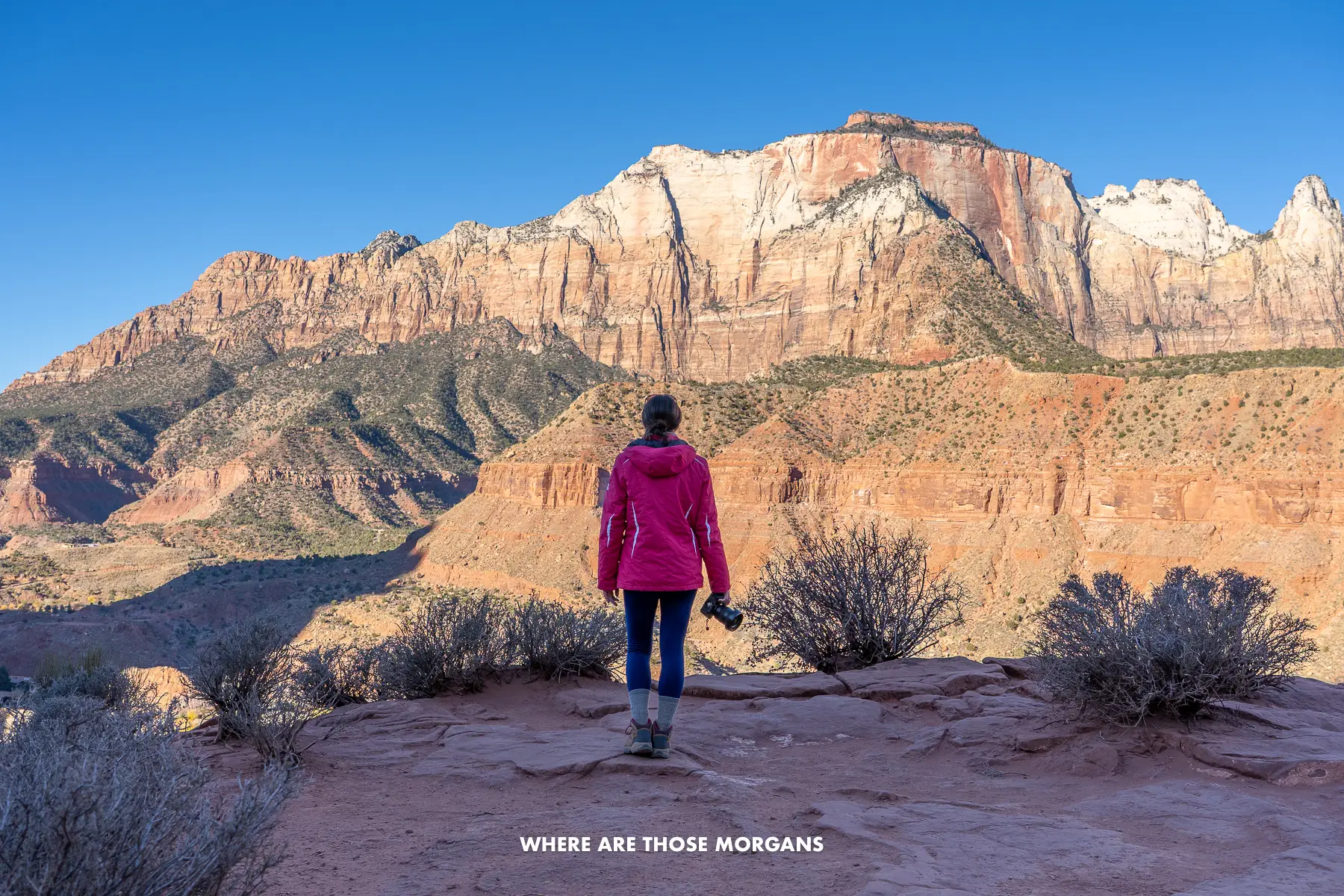 Photo of Kristen walking out to Watchman Trail overlook at sunrise in a coat and holding a camera looking at red rock cliffs lighting up ahead