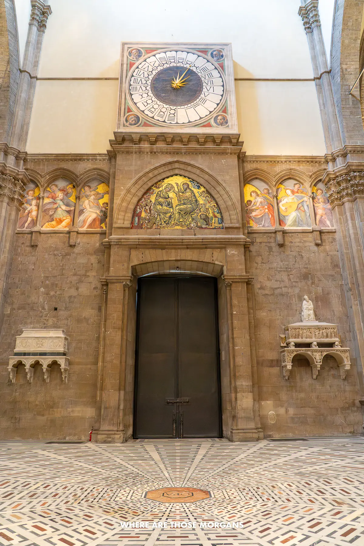 Photo of a 24 hour clock with Roman numerals above a huge door at the entrance to a cathedral with a mosaic floor