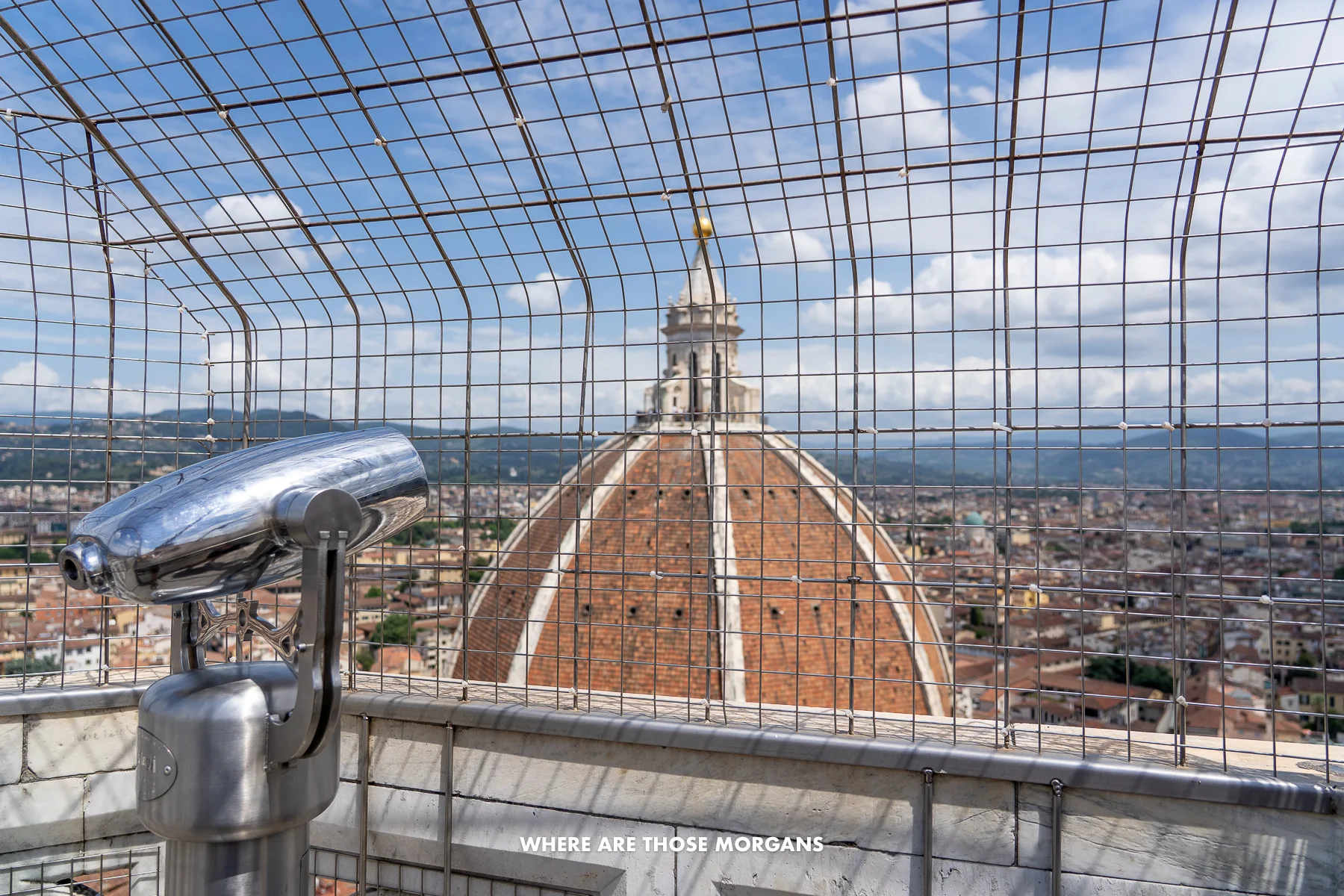 Photo of binoculars looking through a mesh metal fence at the dome of a church in Italy on a sunny day