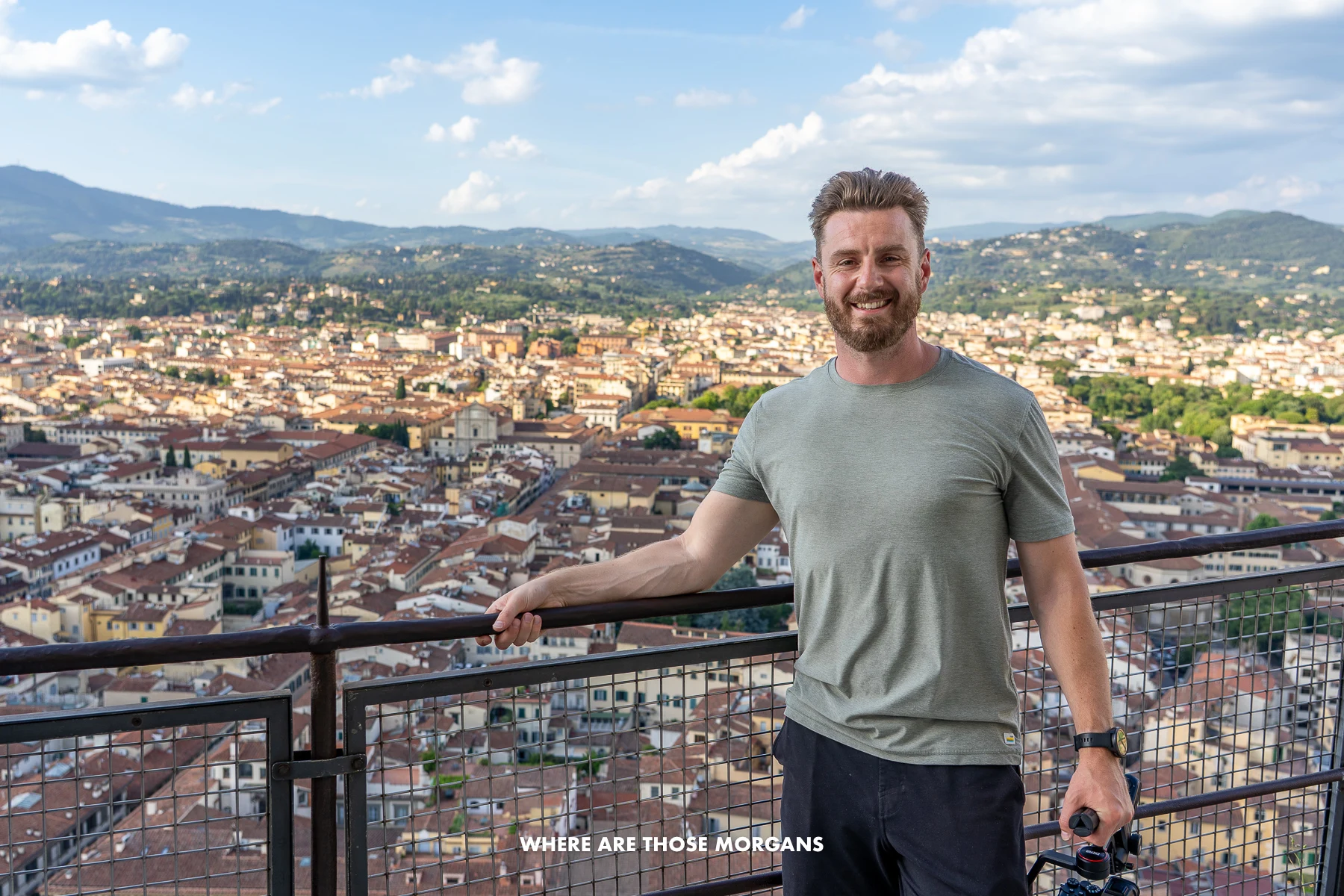 Photo of Mark Morgan from Where Are Those Morgans holding onto a black metal railing with elevated views over the city of Florence Italy behind on a sunny day