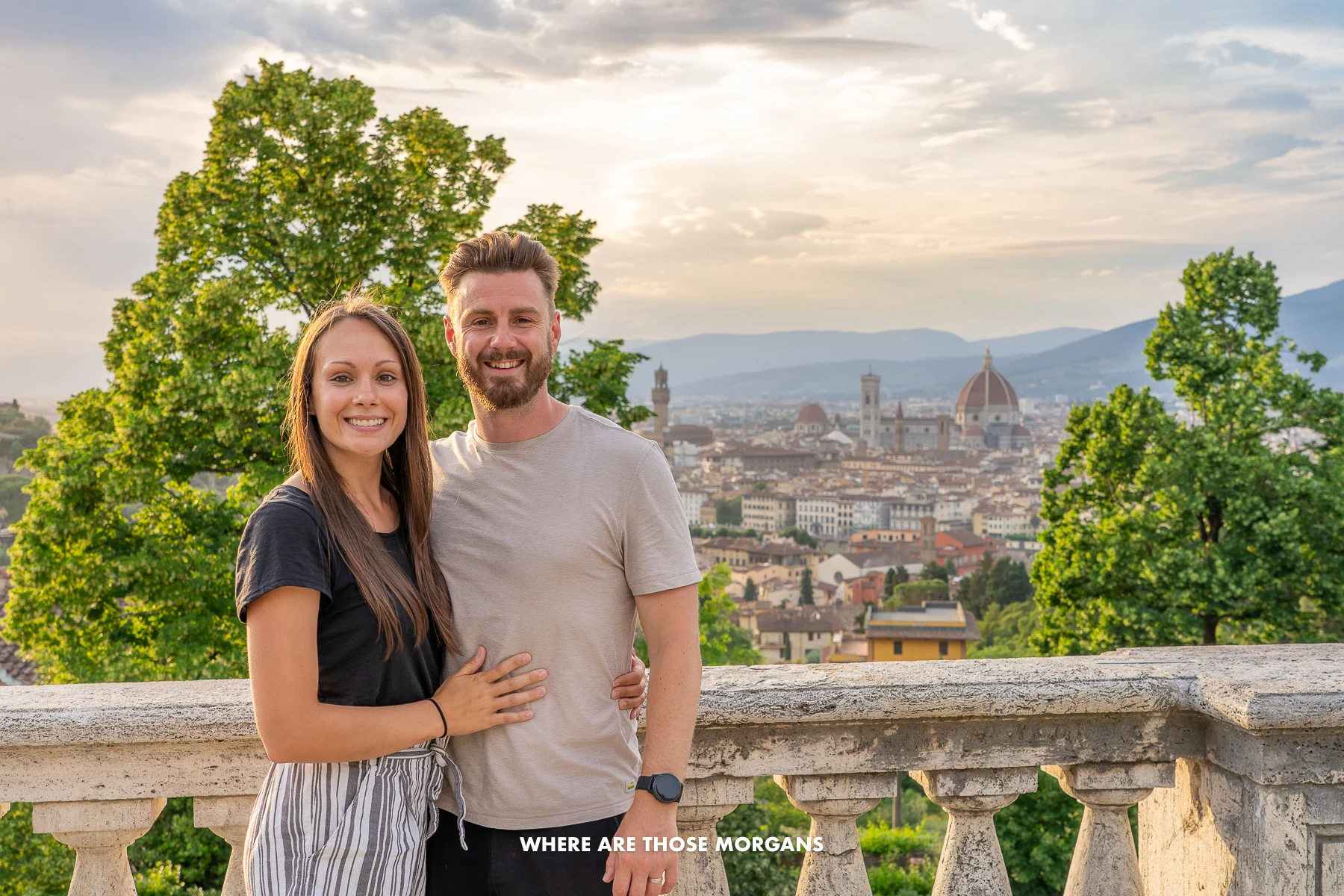 Photo of Mark and Kristen Morgan from Where Are Those Morgans standing together in front of a low stone wall with views of trees and the city of Florence behind at sunset