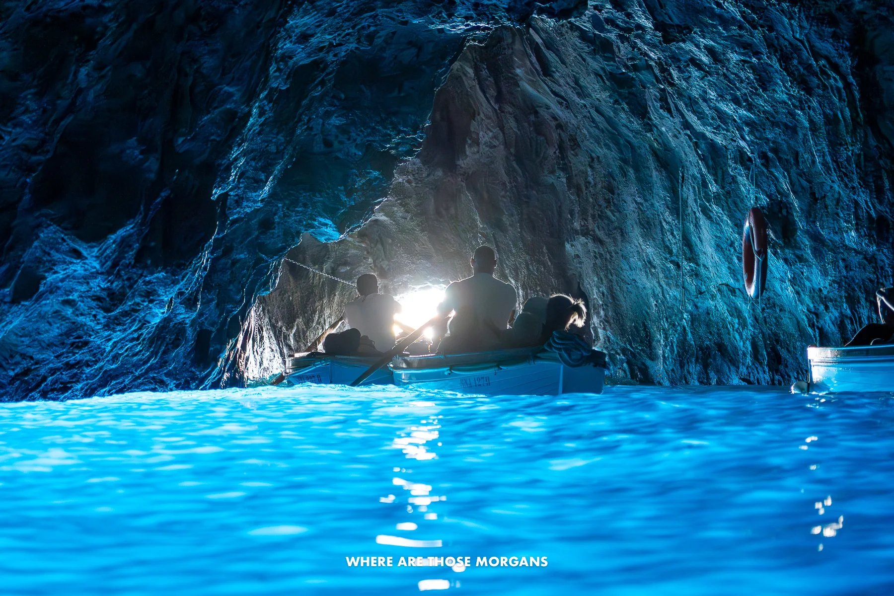 Photo of light blue water inside a small sea cave with a tunnel of light in the distance at the entrance and rowing boats heading for it