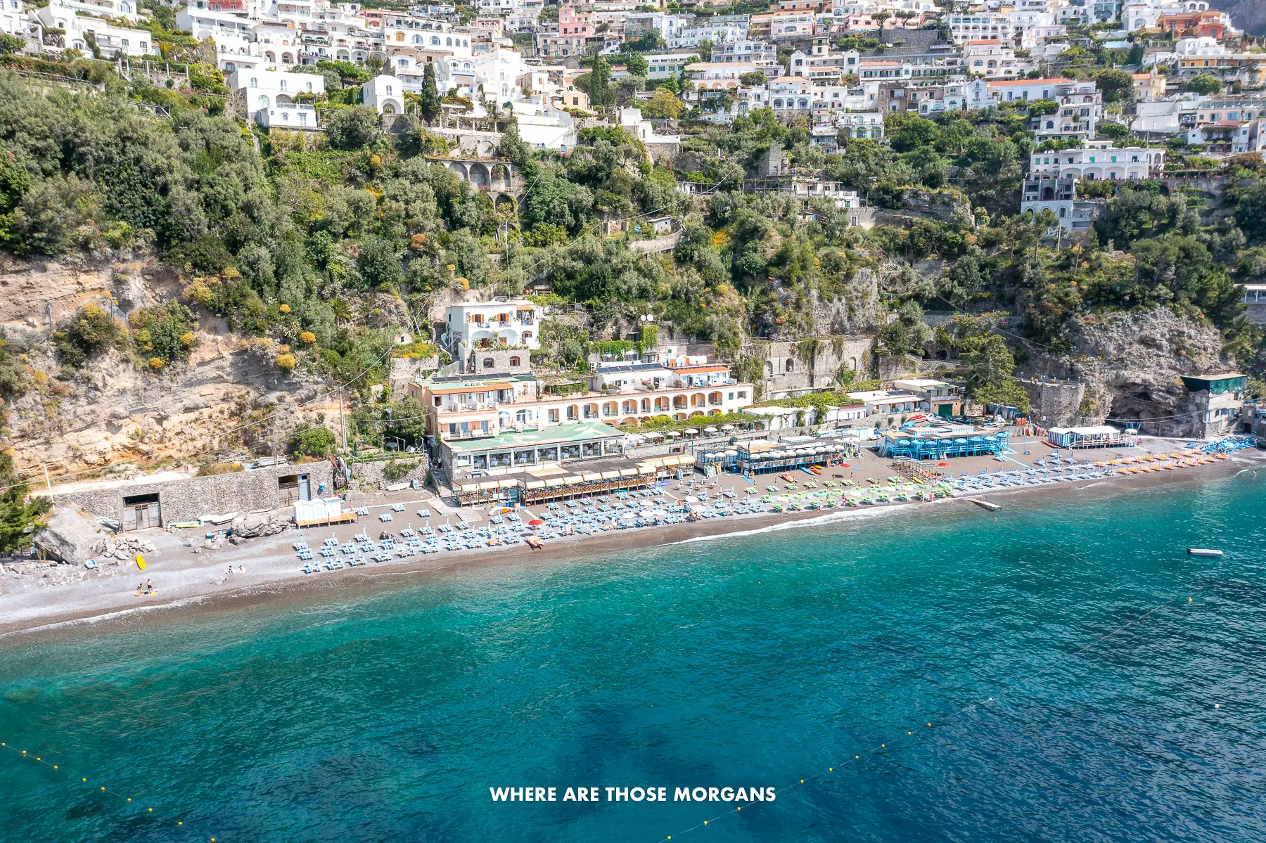 Drone photo of Fornillo Beach in Positano from out at sea with turquoise water leading to the pebble and rock beach, and people sunbathing