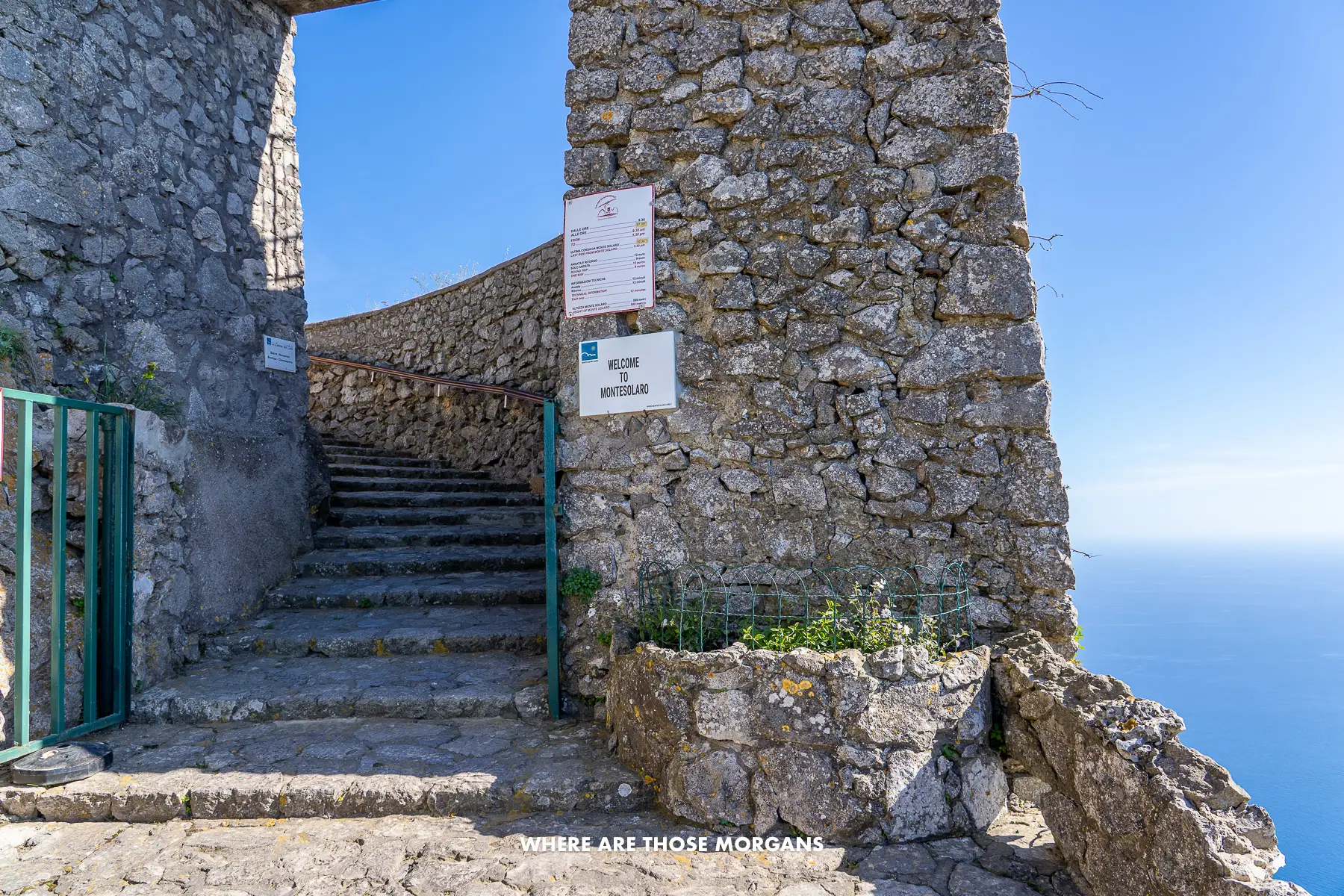 Photo of a stone staircase with a stone arch and a fence to close at the top of a mountain with sea views behind