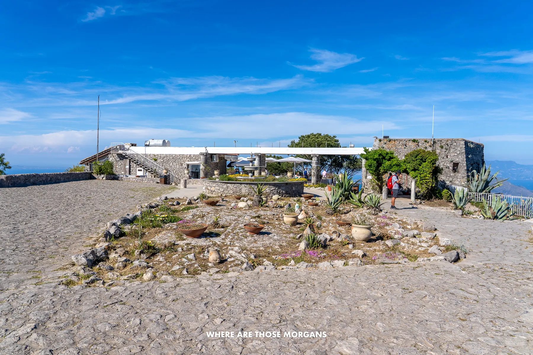 Photo of a flat stone terrace with a small building ahead at the top of a mountain with a deep blue sky and sea views