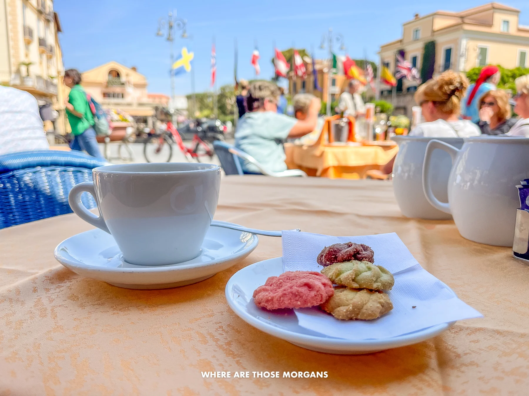 Photo of coffee and biscuits on a table with lots of people in the background of a restaurant on a sunny day