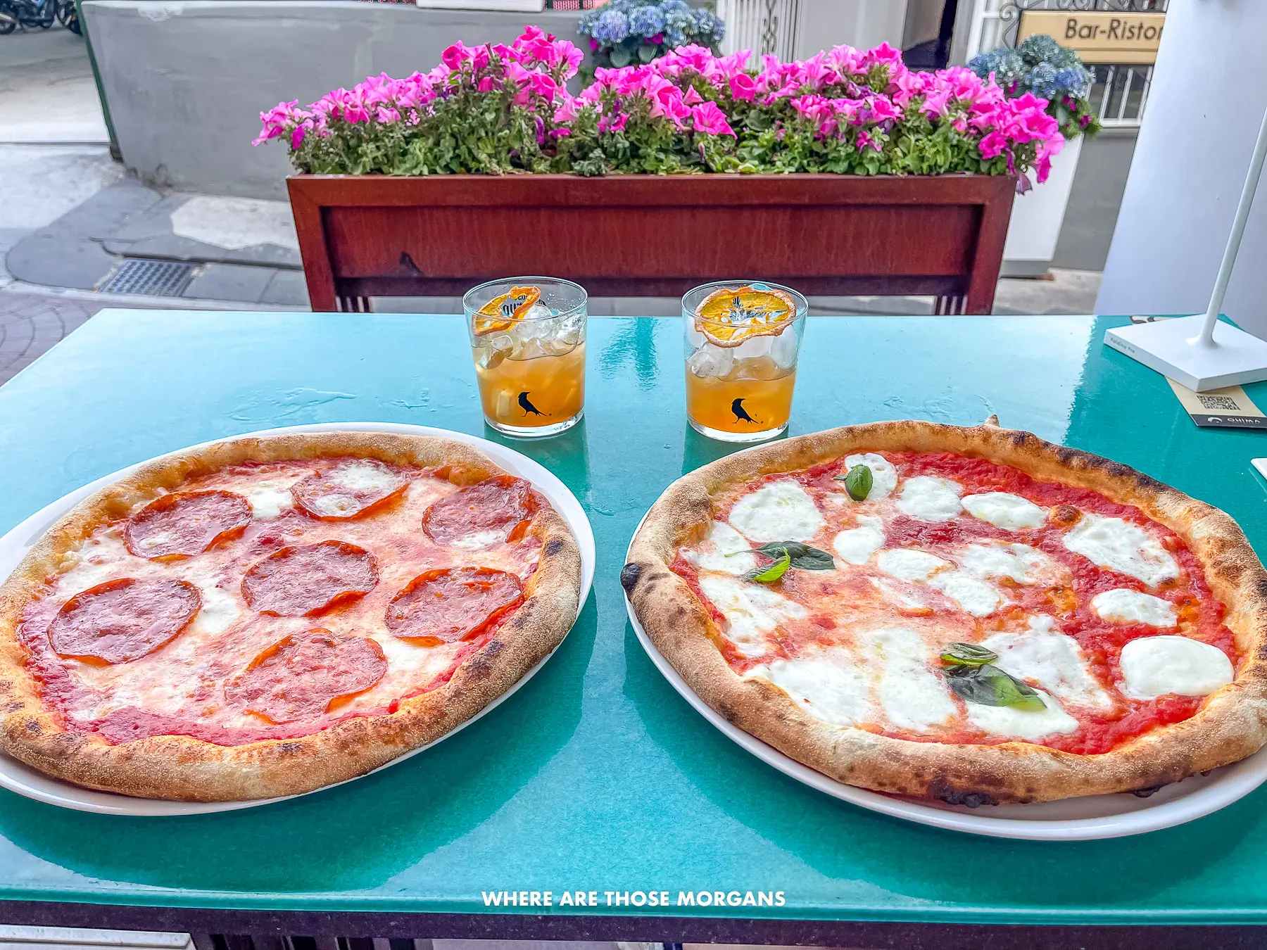 Photo of two Neapolitan pizzas and two amaretto sours on a turquoise table with flowers behind