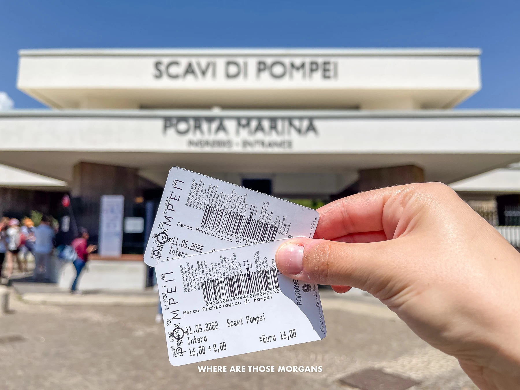 Photo of a hand holding two white attraction tickets with the entrance to Pompeii ruins in the background