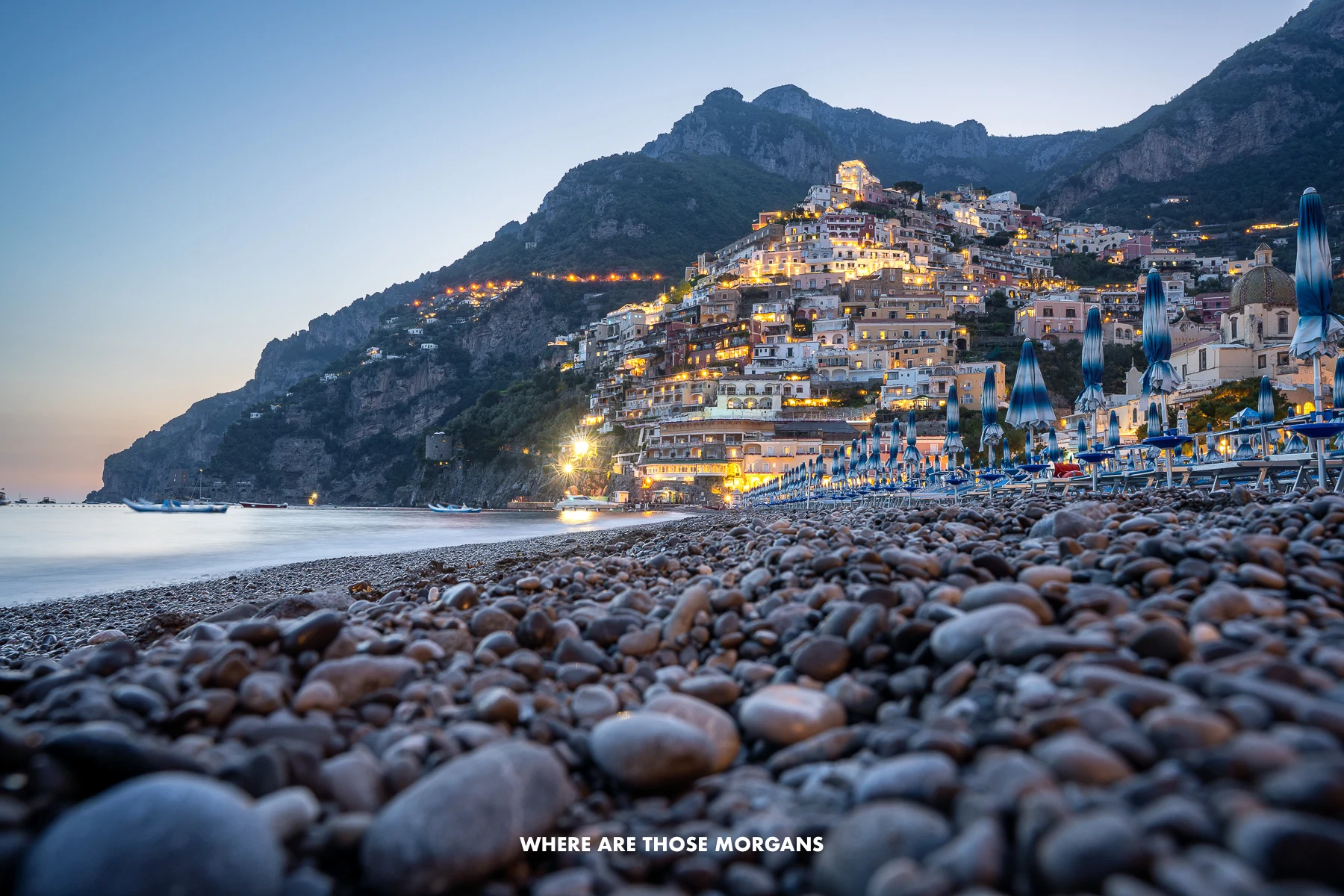 Photo of Positano during twilight after sunset taken right on the beach with close up pebbles and the town lit up in the distance