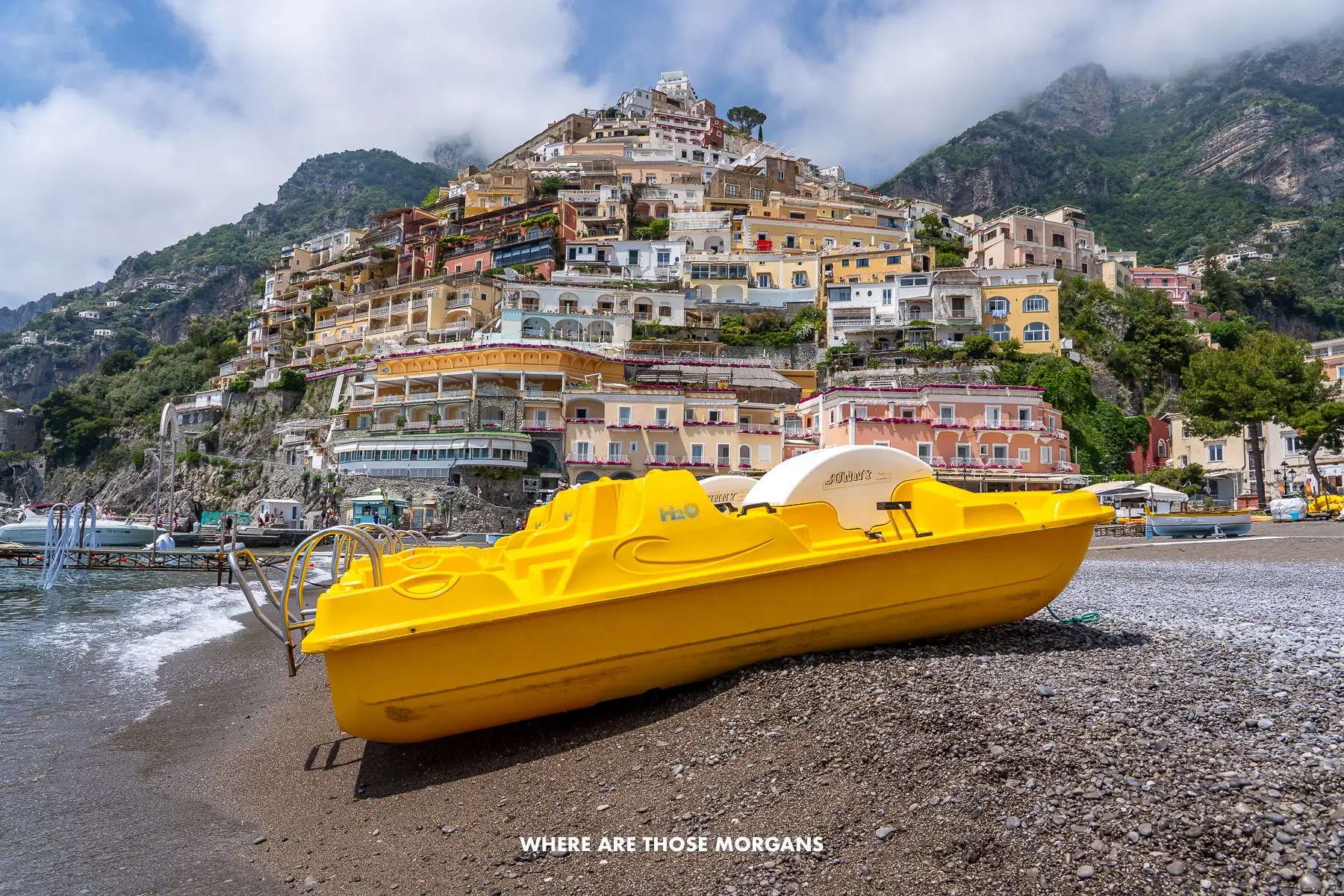 Photo of a yellow pedal boat sat on a pebbly beach with an Amalfi Coast town behind in Italy