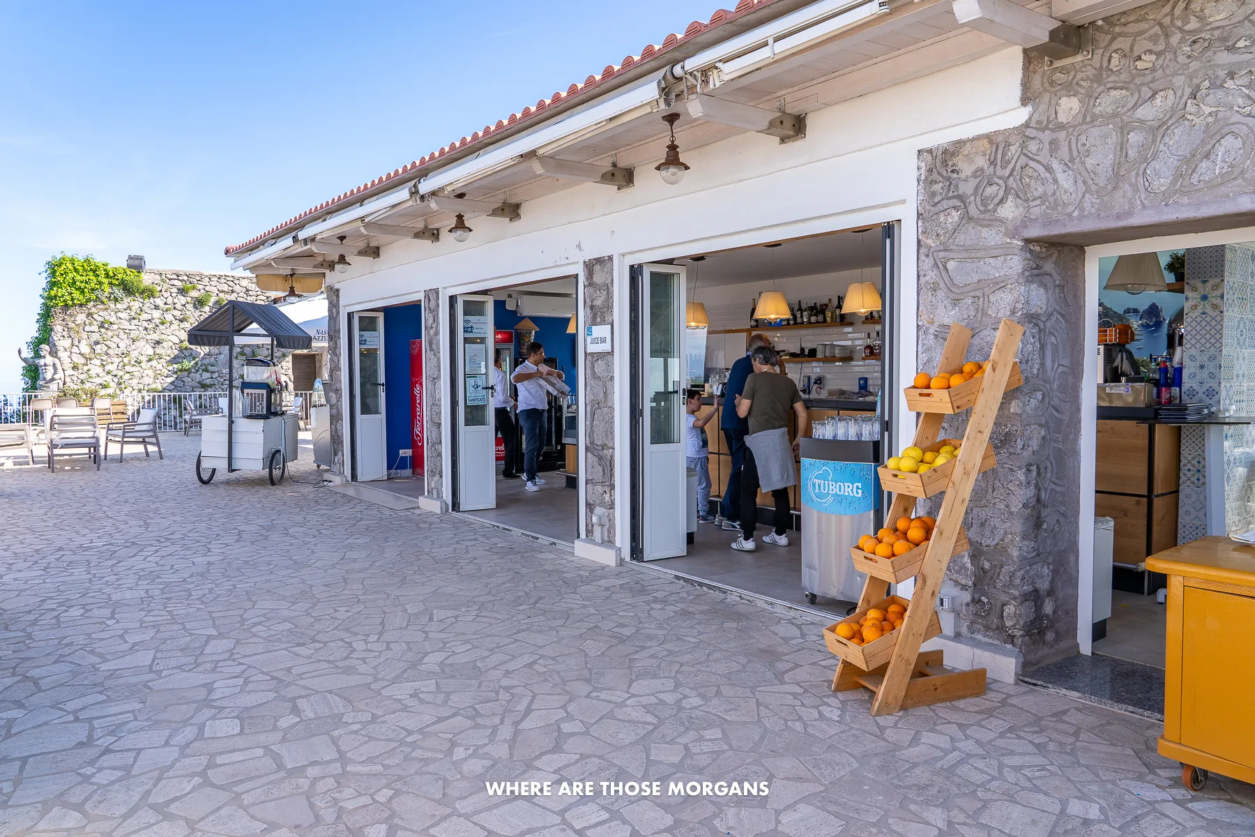 Photo of a small low profile restaurant and cafe with fresh fruit outside and a few people inside