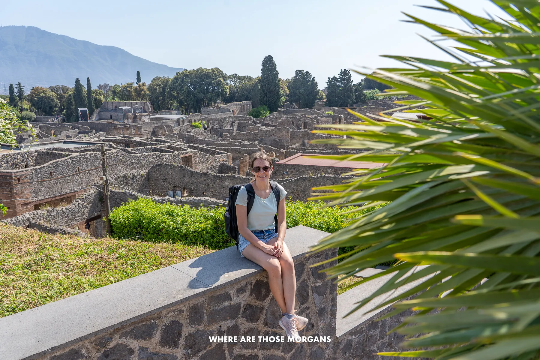 Photo of Kristen from Where Are Those Morgans sat on a wall next to a bush with an ancient ruined town behind in Italy