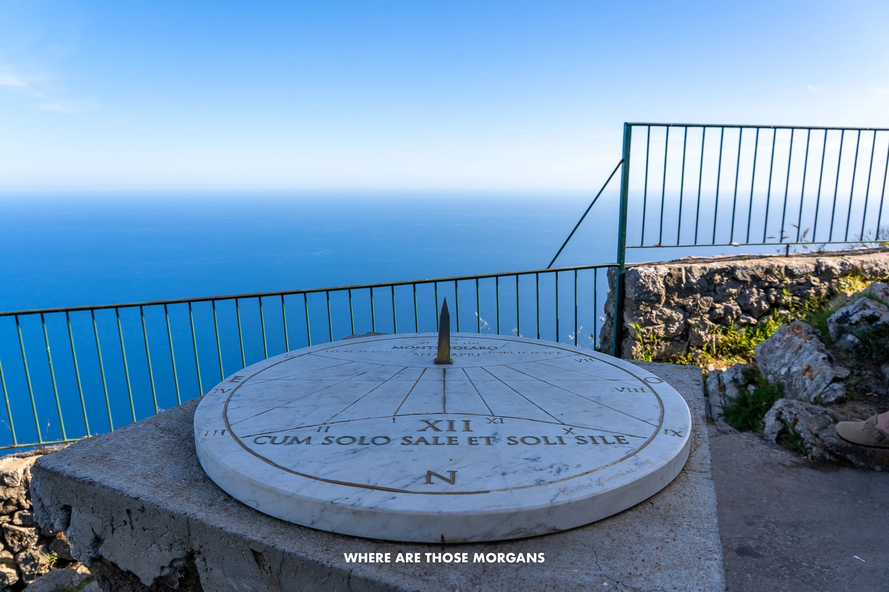 Photo of a sundial in shade with a black fence on a small wall and then nothing but ocean behind