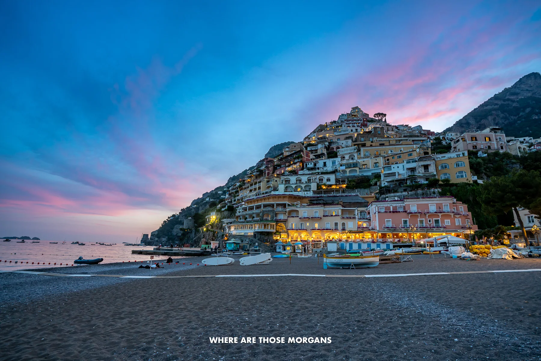 Photo of a beach and Amalfi Coast town on a cliffside backed by colorful skies at sunset