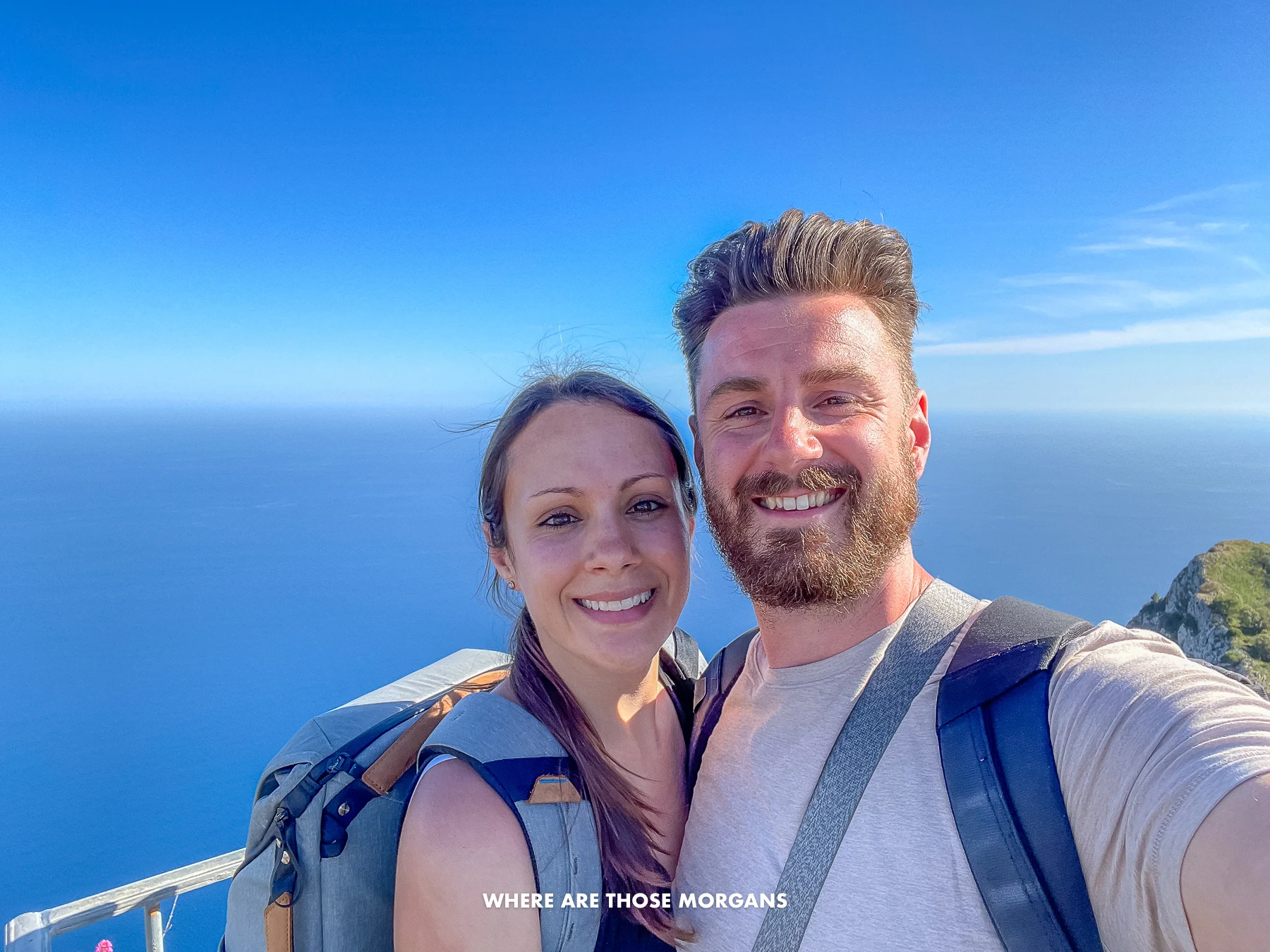 Photo of Mark and Kristen Morgan from Where Are Those Morgans taking a selfie at the top of Monte Solaro in Capri with blue sky and sea behind on a sunny day