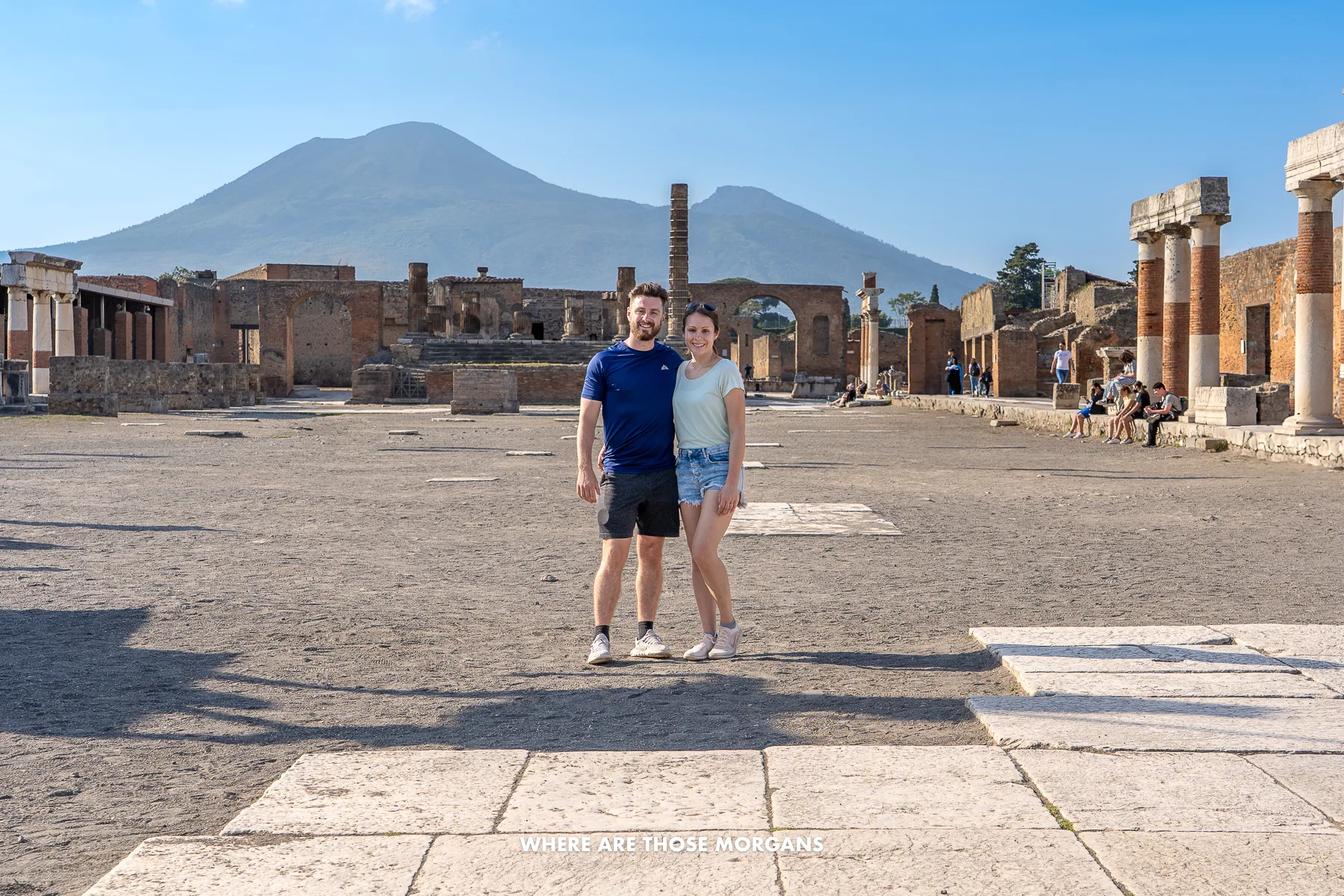Photo of Mark and Kristen Morgan standing together in the middle of the Forum of Pompeii with Vesuvius in the background and tourists exploring the ruins on a sunny day in Italy