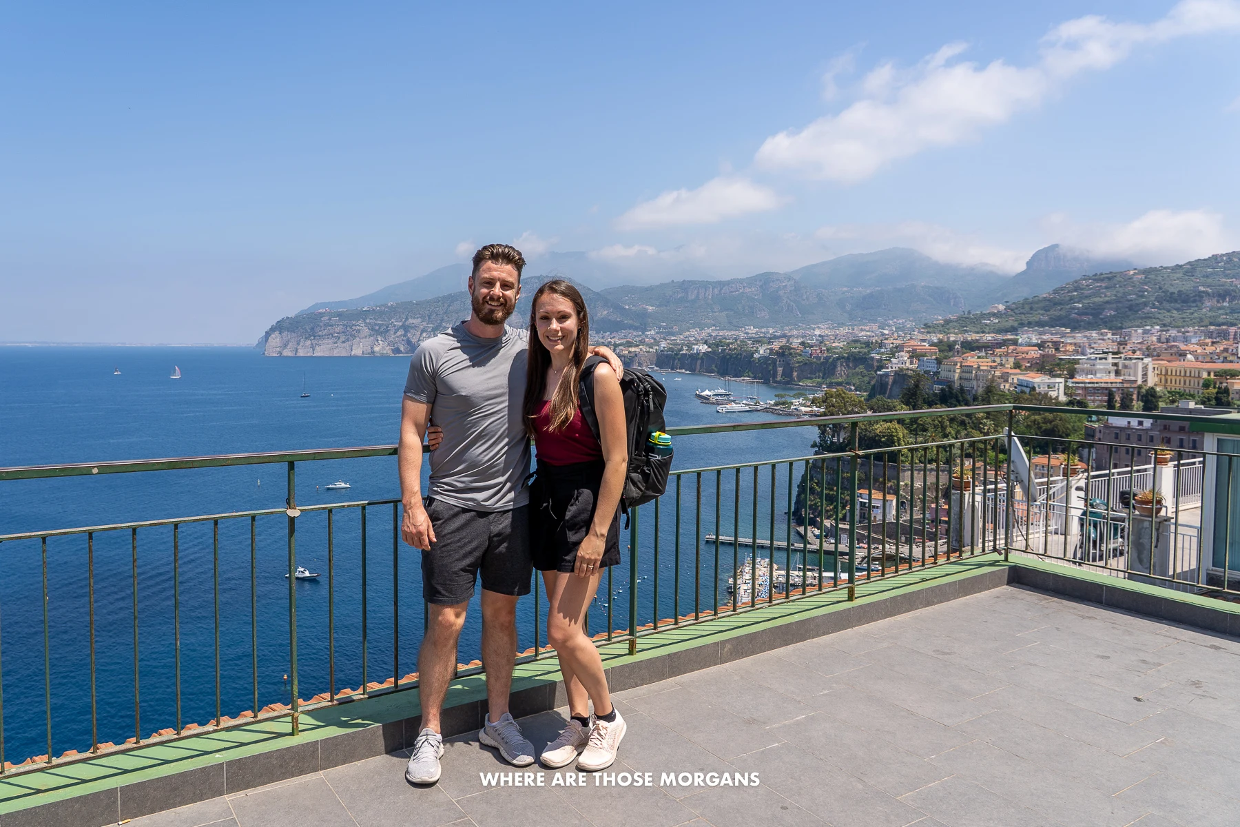 Photo of Mark and Kristen Morgan from Where Are Those Morgans standing together on an elevated concrete balcony on a warm day with views over Sorrento, Italy and the Gulf of Naples behind