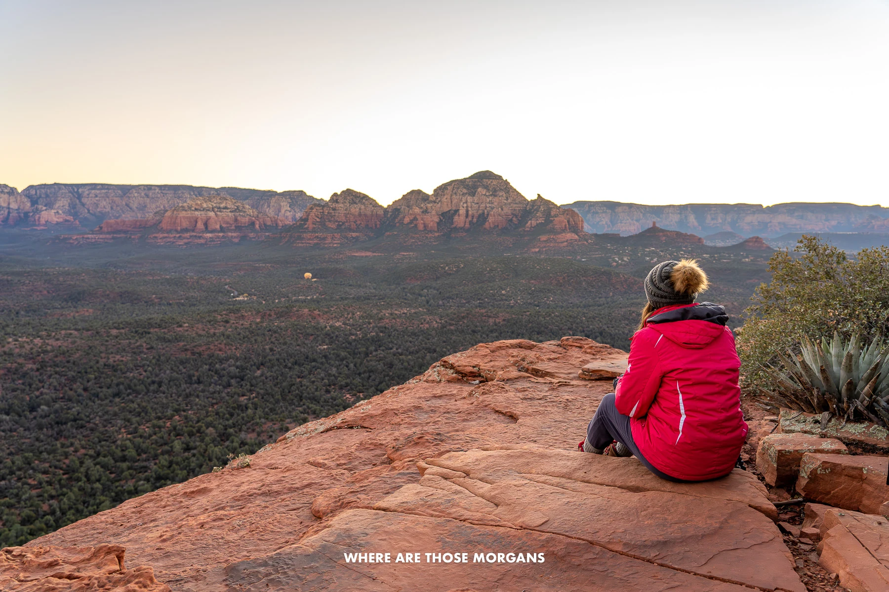 Photo of Kristen Morgan sat in a red coat and wooly hat on red rocks looking at a sunrise view over a valley and red cliffs in Sedona