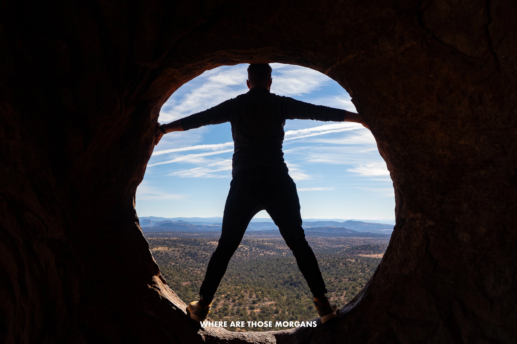 Photo of Mark Morgan standing inside a window of a cave filled with shadows silhouetted against the bright outdoor landscape