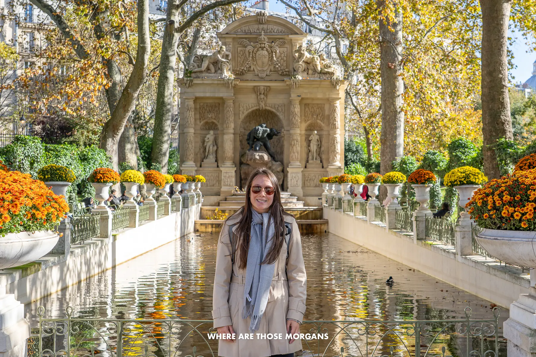 Photo of Kristen Morgan standing in a coat and sunglasses in front of a fountain inside a park with flowers on either side of the water