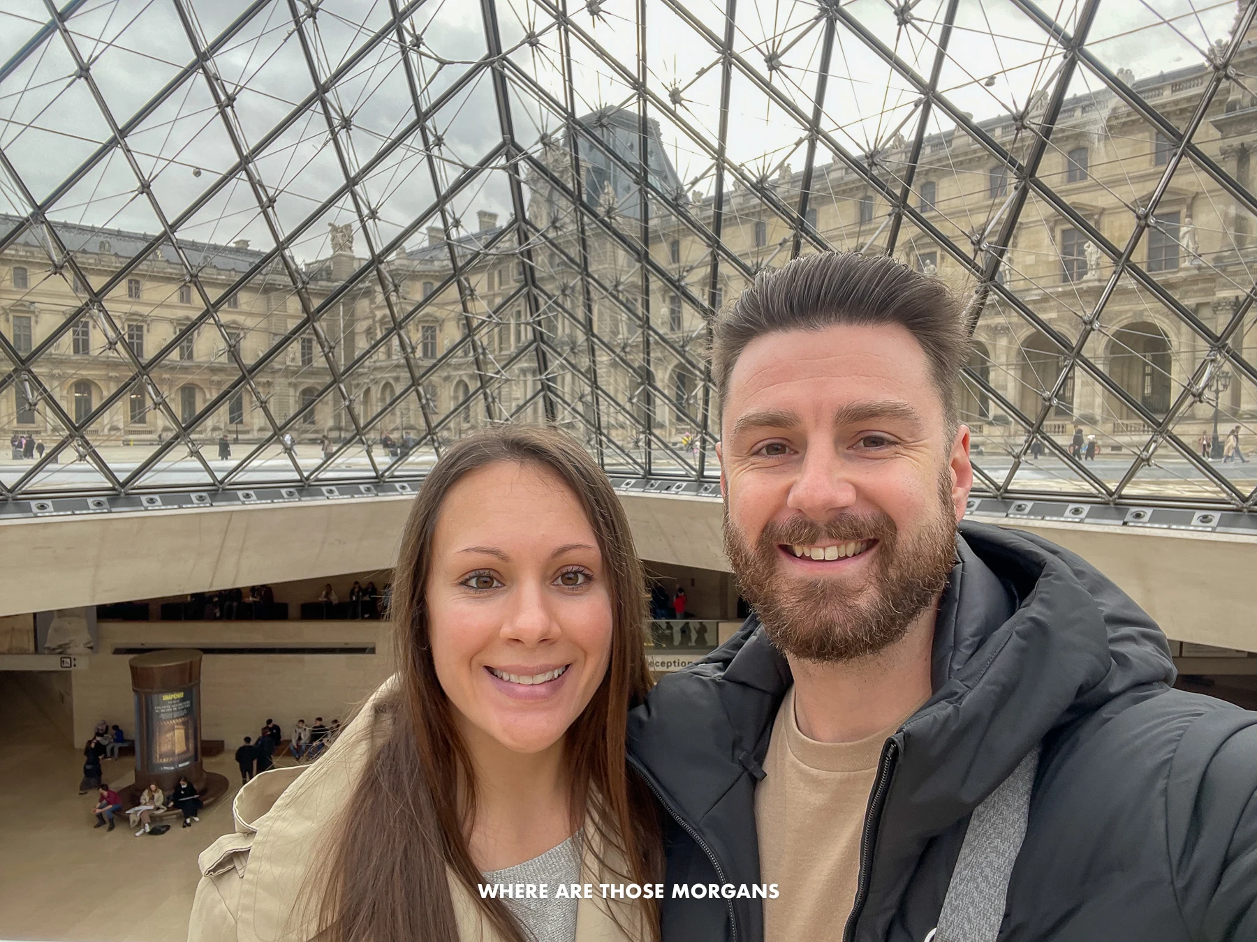 Photo of Mark and Kristen from Where Are Those Morgans taking a selfie at the entrance to the Louvre in Paris with coats on and the large glass pyramid above