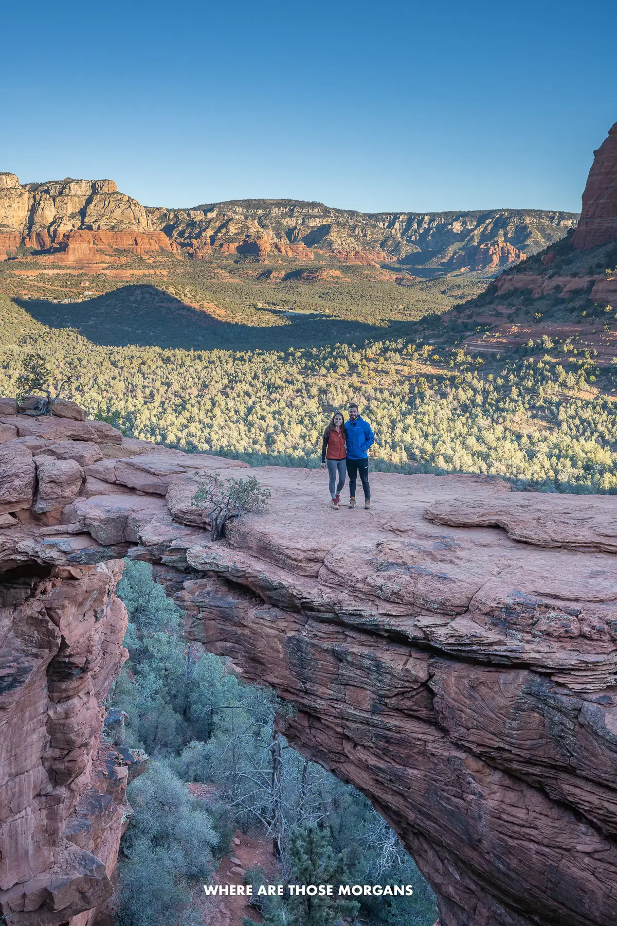 Photo of Mark and Kristen from Where Are Those Morgans standing together on a natural red rock arch with views over a valley covered in trees and distant red rocks behind at sunrise