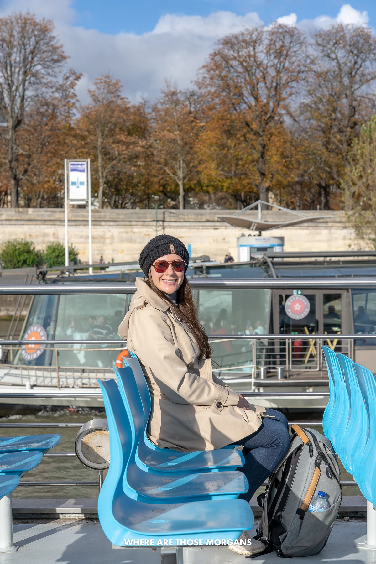 Photo of Kristen Morgan sat on blue plastic seats on a Seine river cruise in Paris with a coat and hat on a sunny day