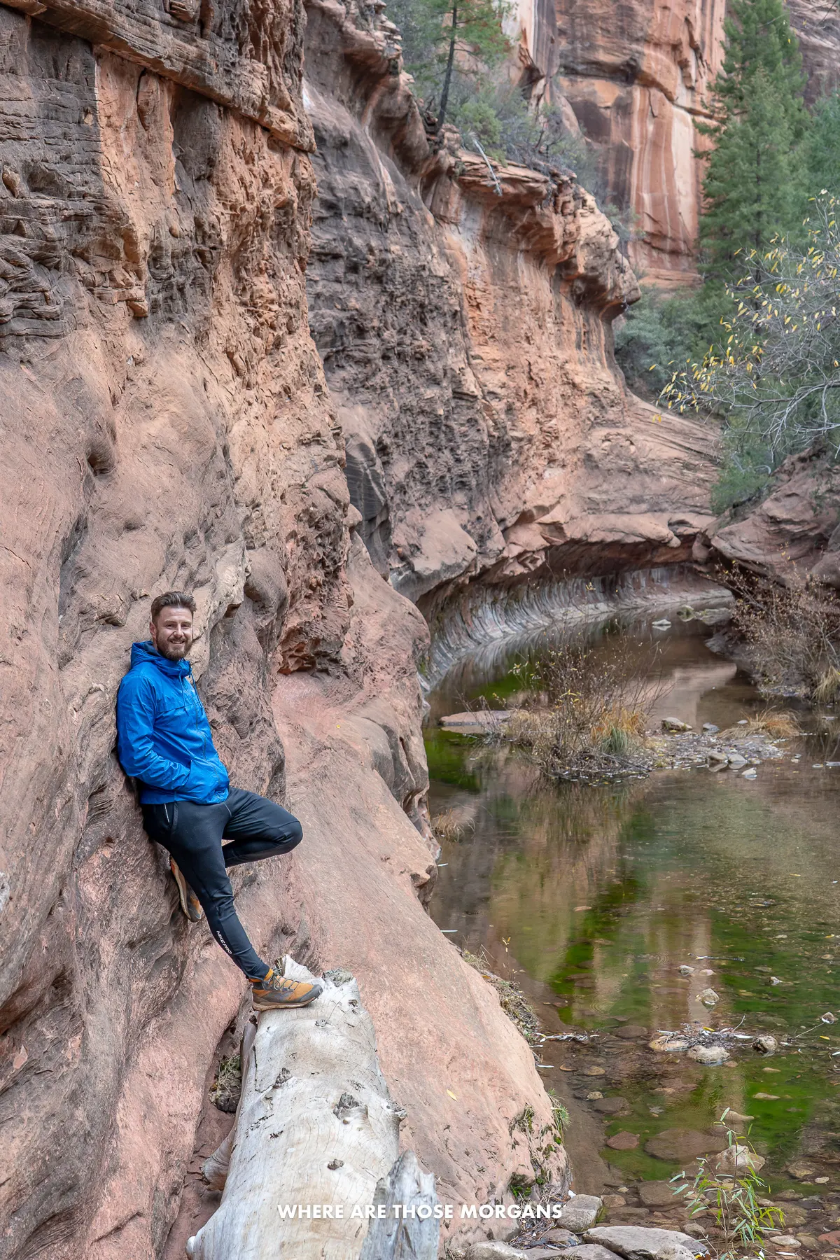 Photo of Mark Morgan leaning up against a red rock wall next to a river running through a subway-shaped formation