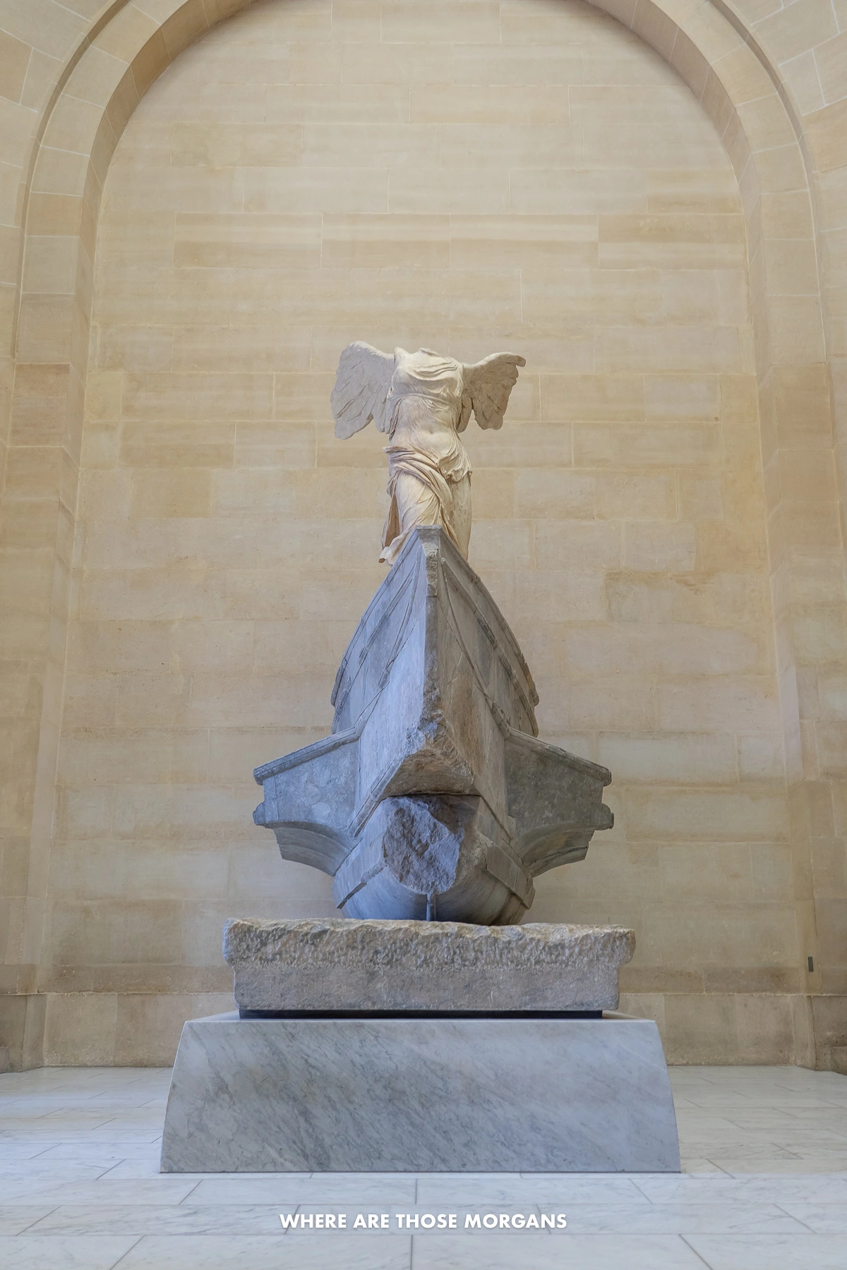 Photo of a sculpture with wings and missing a head backed by a marble wall with arch inside a museum