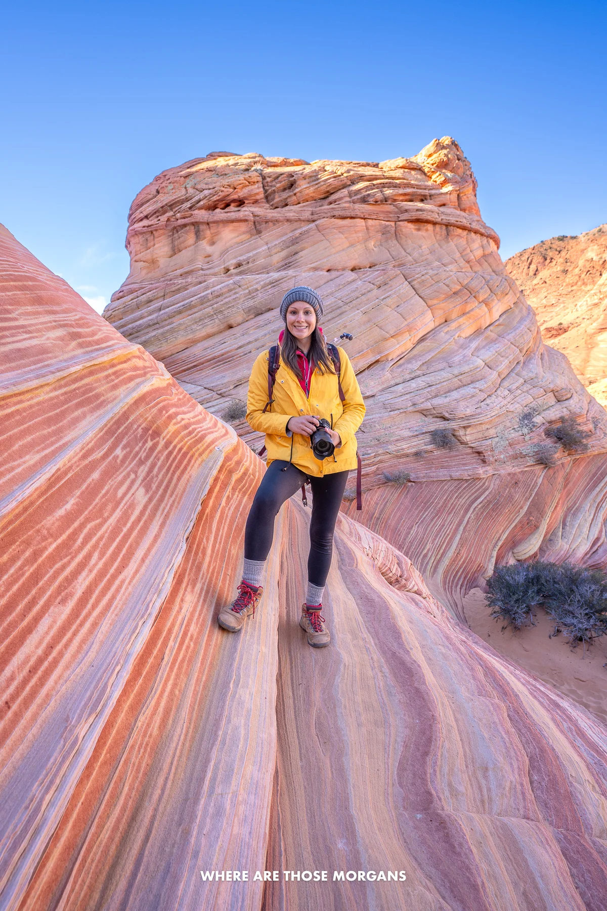 kristen morgan the wave arizona Hiker holding camera in a yellow coat and wooly hat standing on a sloped colorful rock formation with a clear blue sky