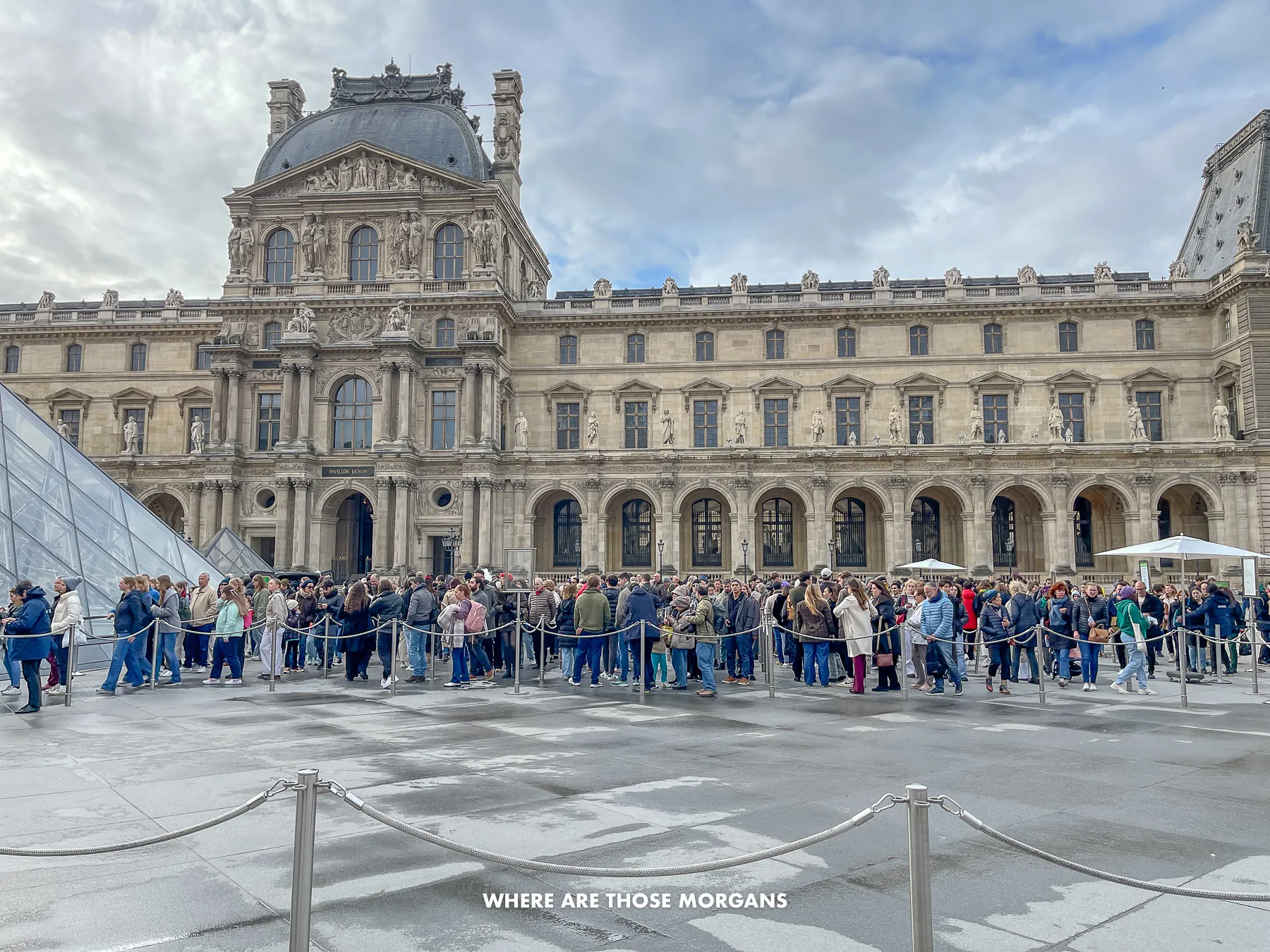 Photo of a long line of tourists waiting in a courtyard to get inside the Louvre Museum