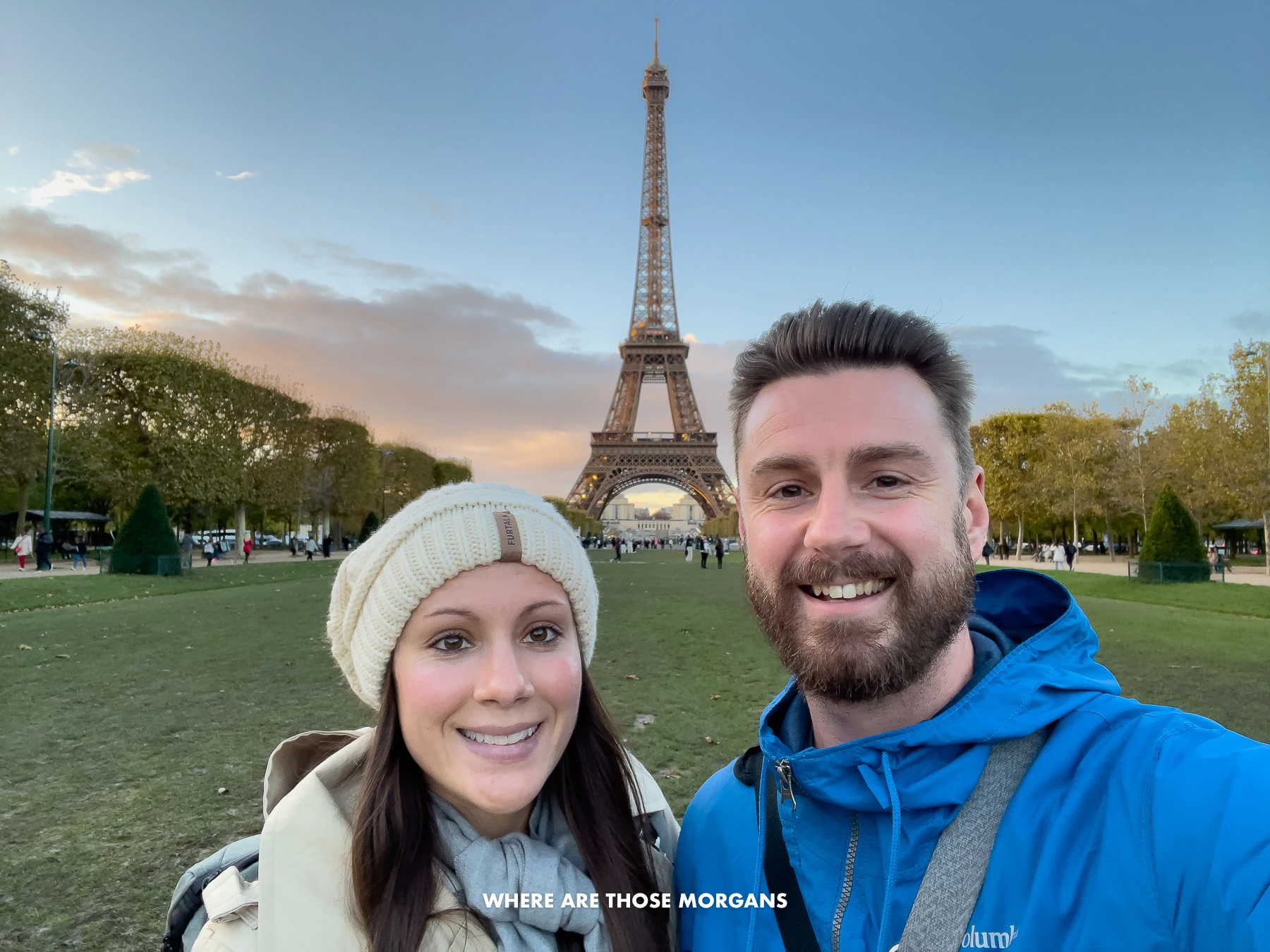 Mark and Kristen from Where Are Those Morgans taking a selfie in coats and hats in November 2023 at sunset on the Champ du Mars with the Eiffel Tower in the background and colors in clouds in the sky