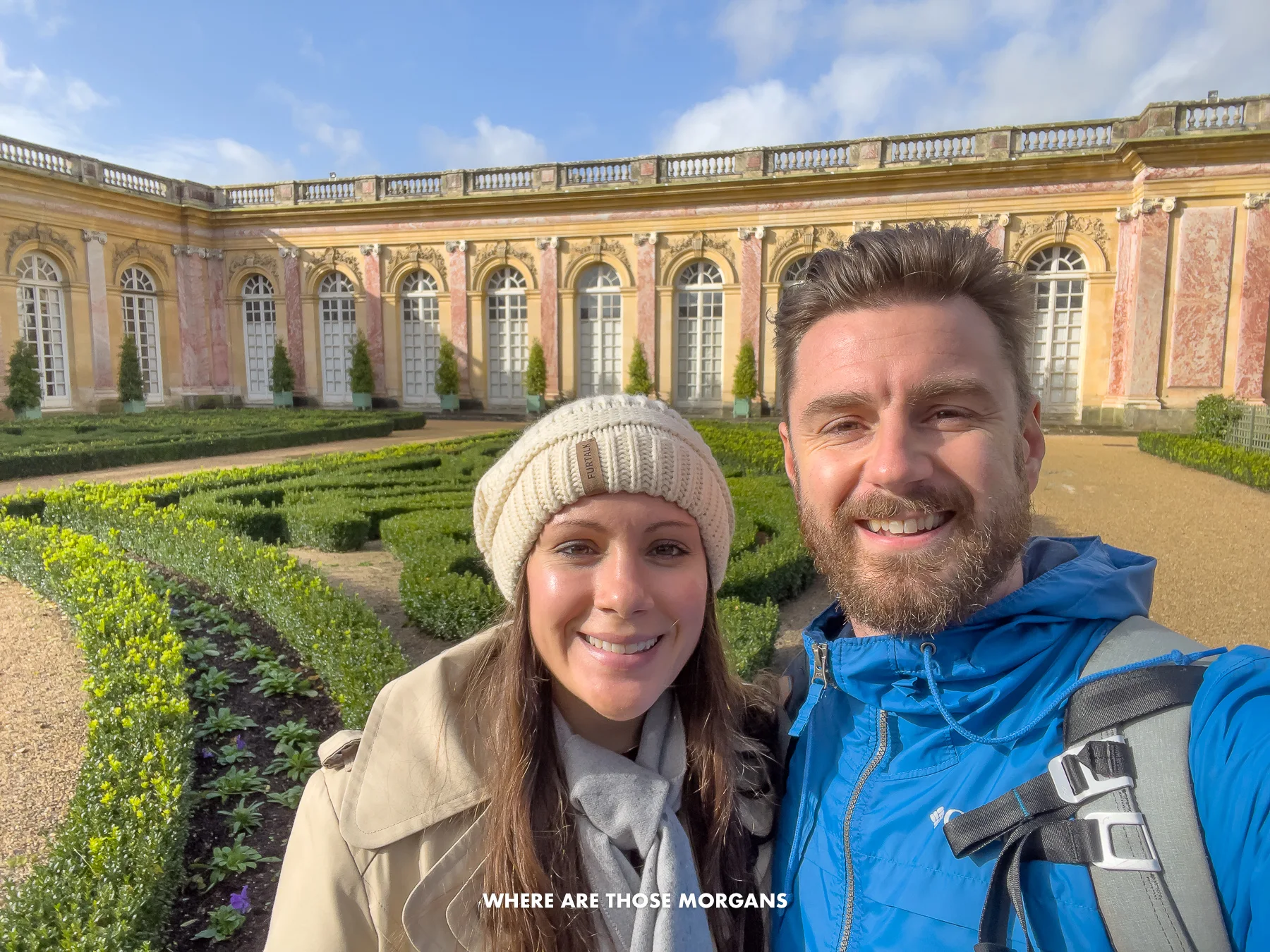 Mark and Kristen from Where Are Those Morgans taking a selfie in the gardens of the Trianon Estate in the Palace of Versailles grounds wearing coats on a sunny day with manicured bushes and an old building behind