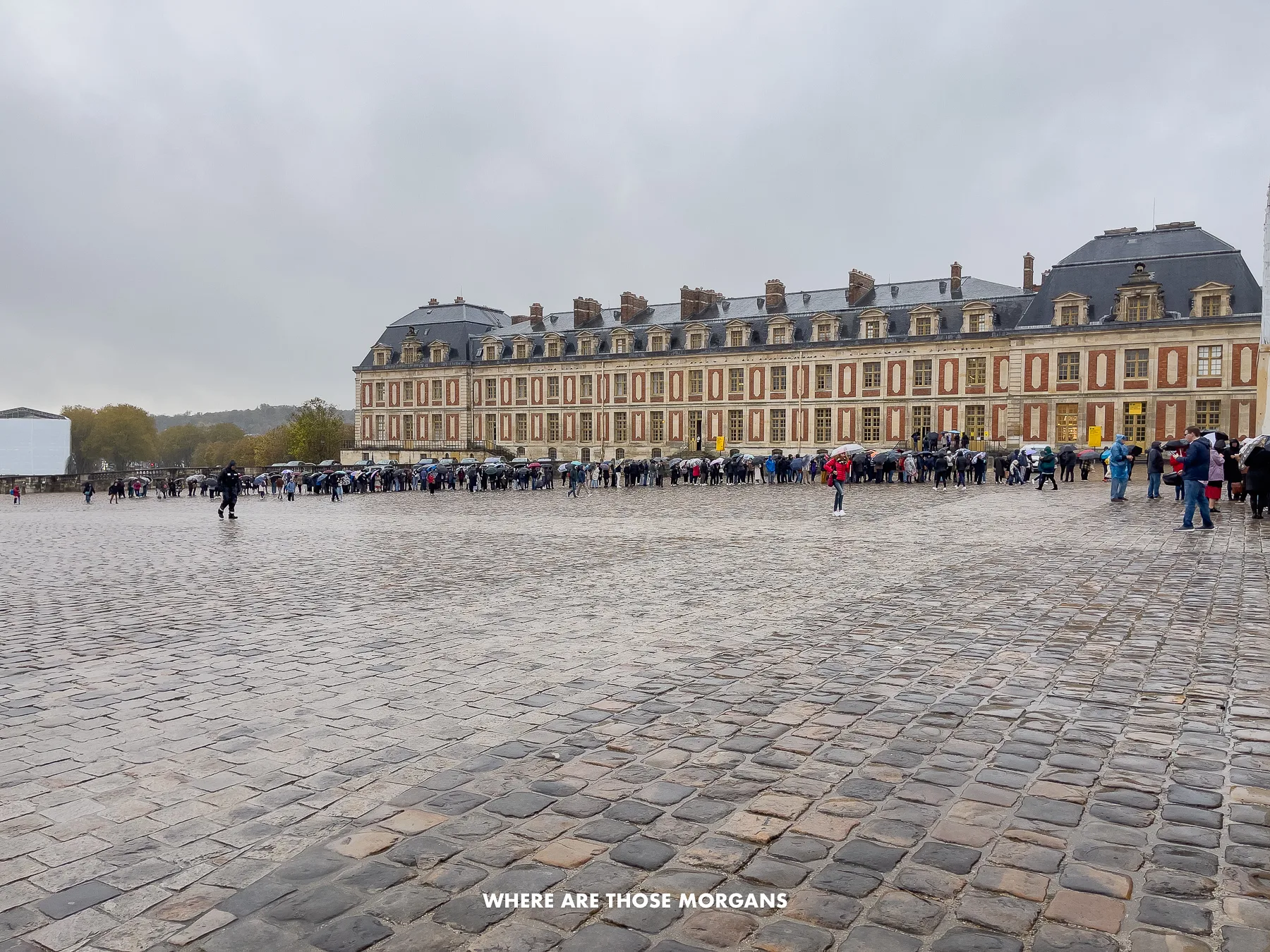 Vast cobbled plaza covered in rain with a long line of tourists wearing waterproofs waiting to get inside grand buildings in France