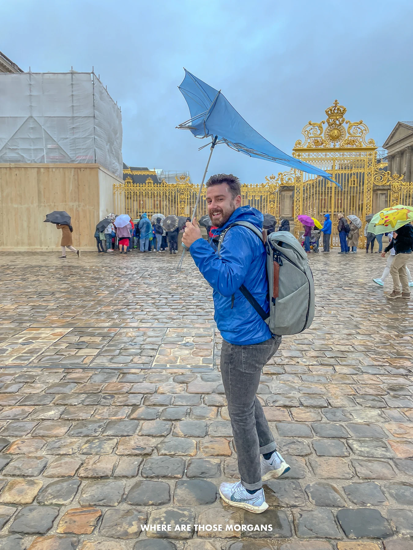 Mark from Where Are Those Morgans laughing and walking on a cobblestone plaza in a coat holding an umbrella that has turned inside out due to strong wind