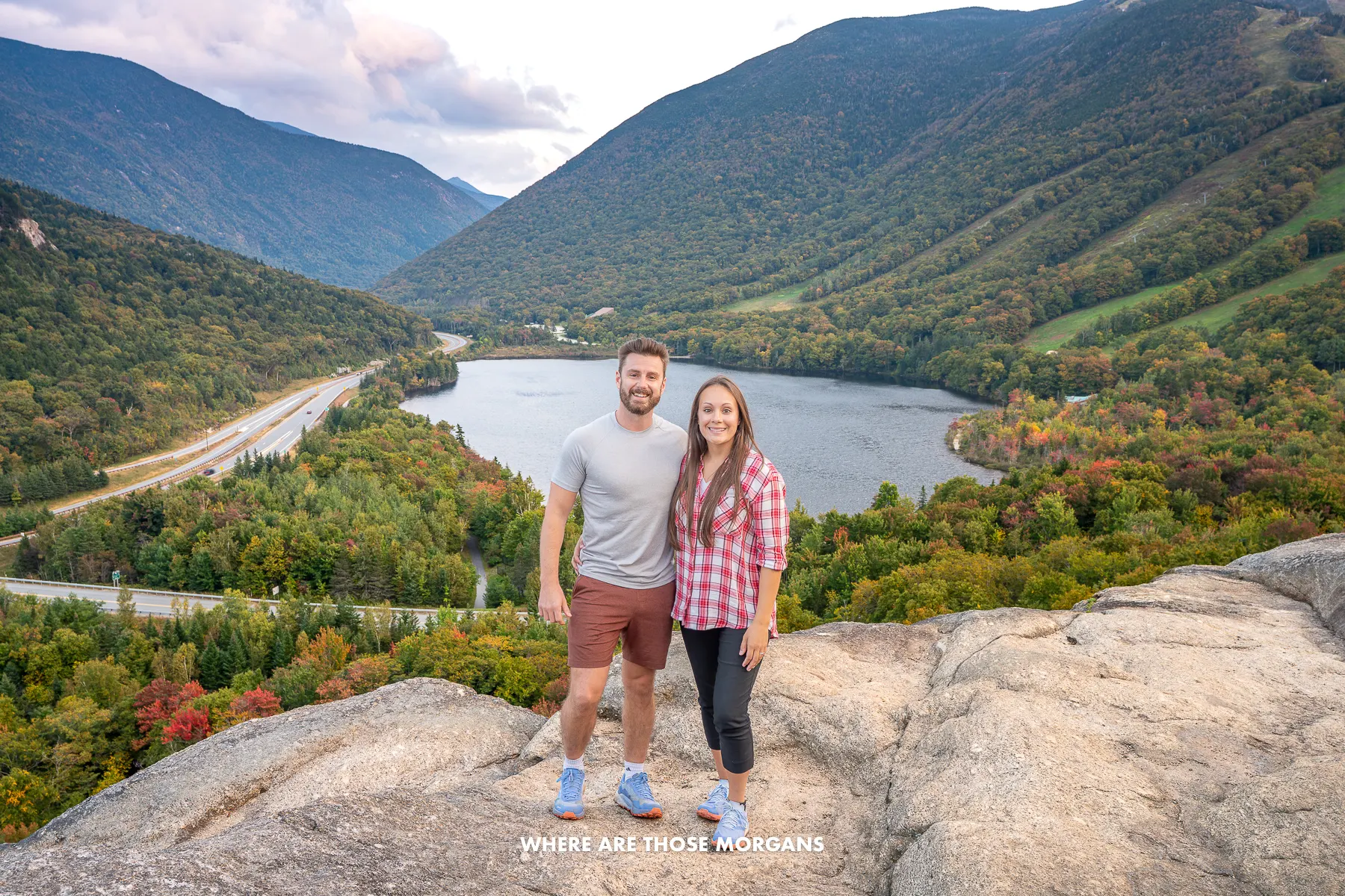 Photo of a couple standing together on a rocky hiking trail summit with views over a lake and rolling hills covered in green tree leaves just as the foliage is turning in fall