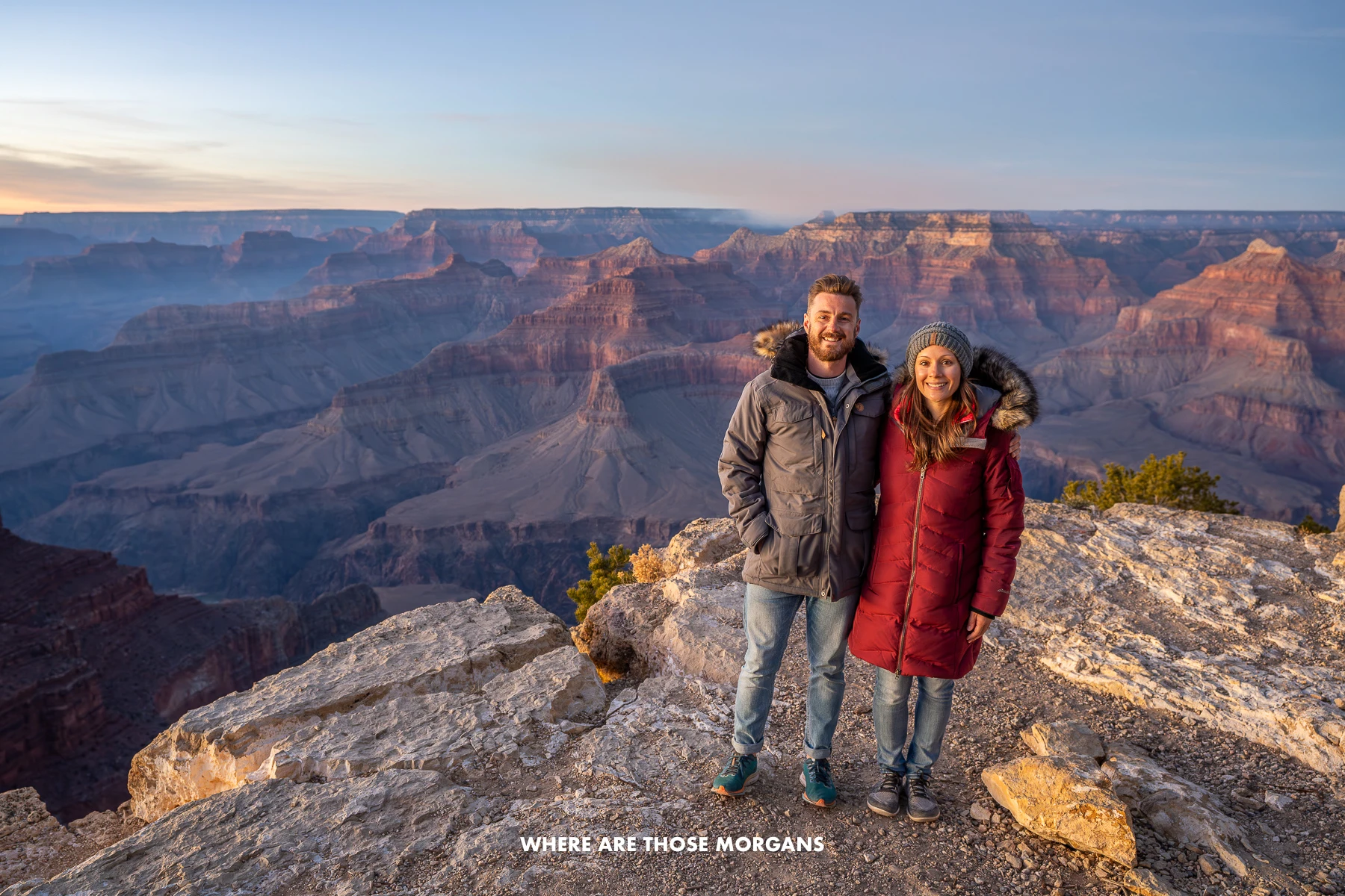 Photo of a couple standing together in winter coats on a rocky plateau with views over a deep canyon at sunset