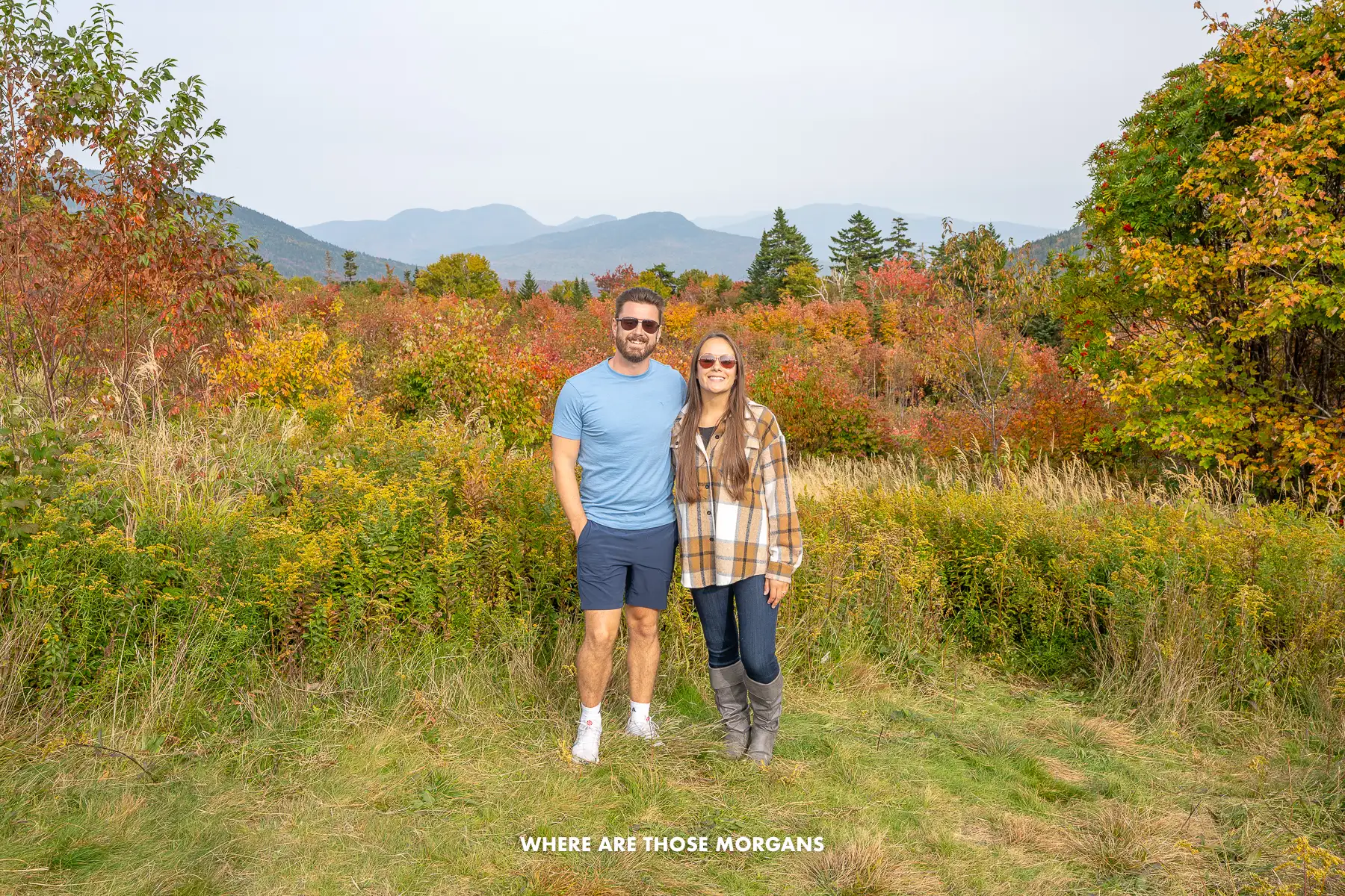 Photo of a couple standing together for a photo in front of fall foliage vegetation and distant rolling hills