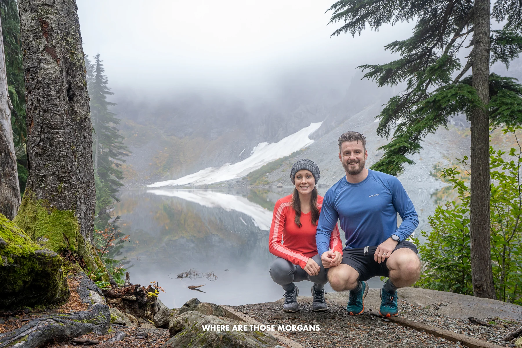 Photo of two hikers crouching next to each other in a forest hiking area with a lake behind on a cloudy day