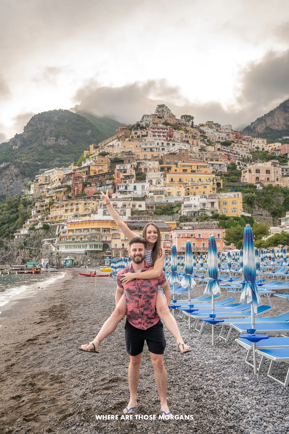Photo of a couple on a gravel beach next to blue sun loungers with a small town built into a hill on a cloudy but sunny day