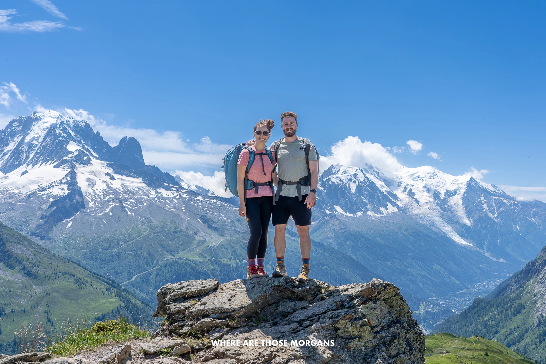 Photo of Mark and Kristen Morgan from Where Are Those Morgans standing together on a large rock in the Alps with hiking gear and backpacks backed by rugged snow capped mountains on a sunny day with clear blue sky