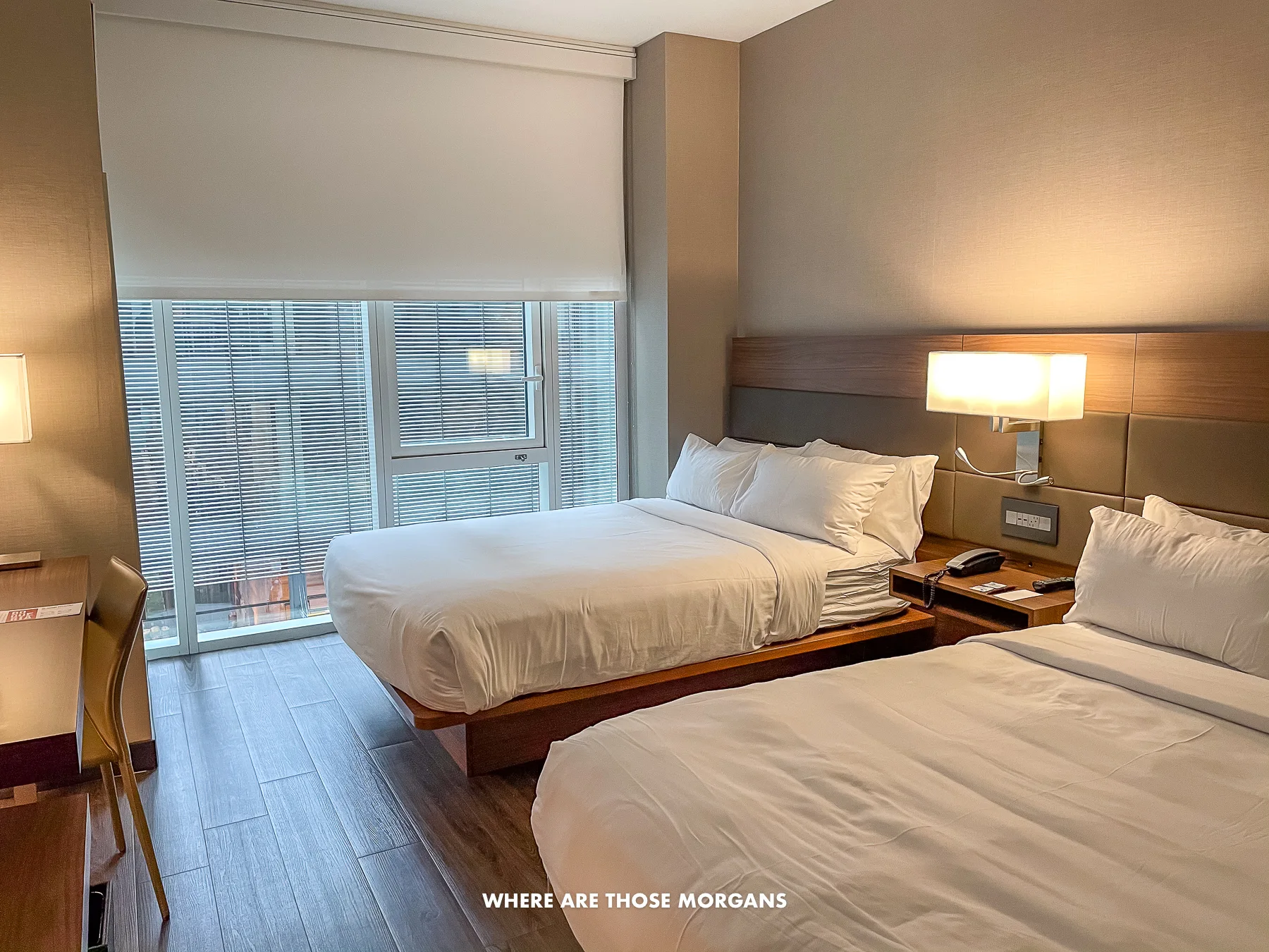Hotel room in NYC with two queen beds and white sheets against a wooden floor with a light on and shades half drawn