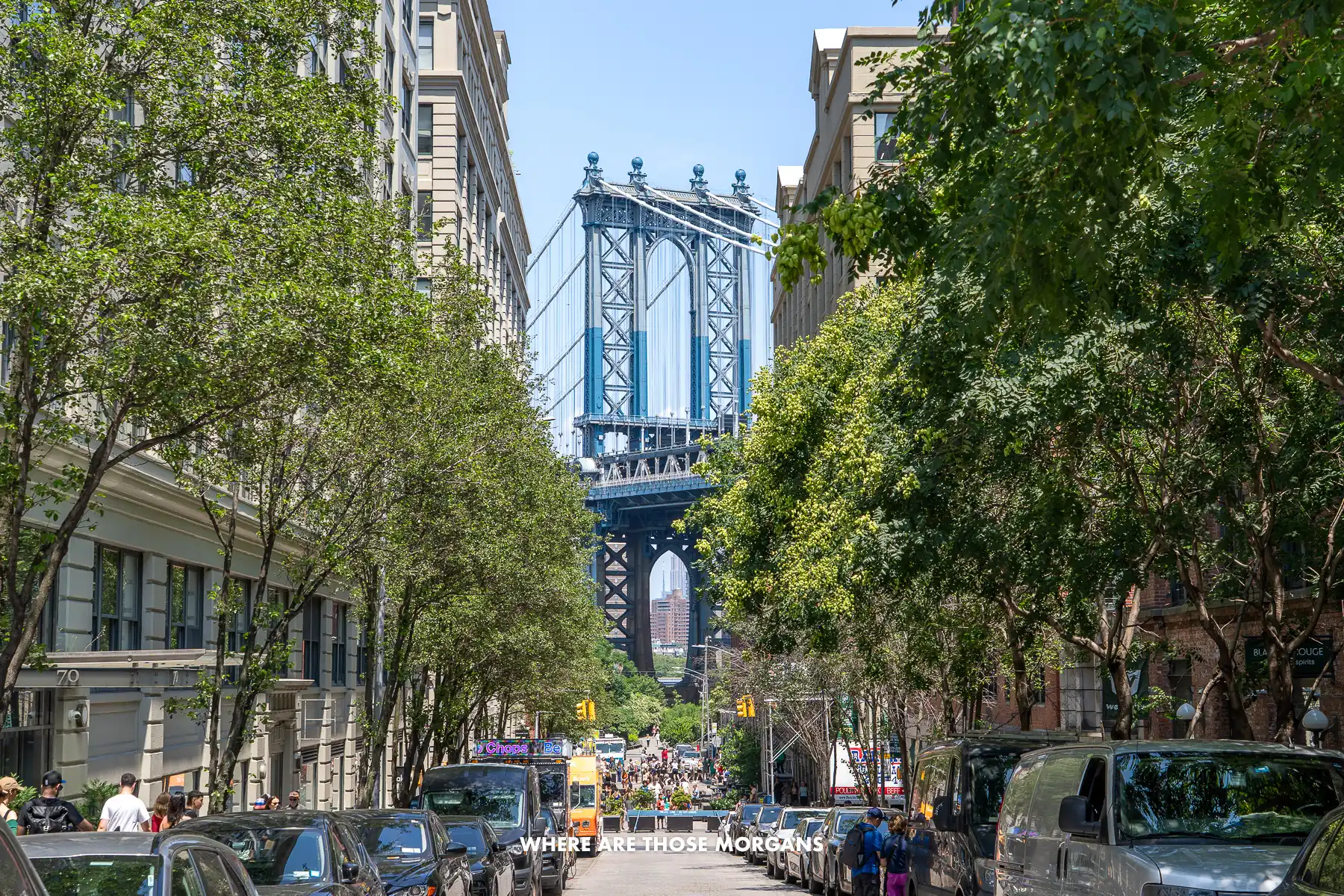 View looking down the tree lined Washington Street between brick buildings with the massive blue Manhattan Bridge bursting into the sky at the end in DUMBO Brooklyn