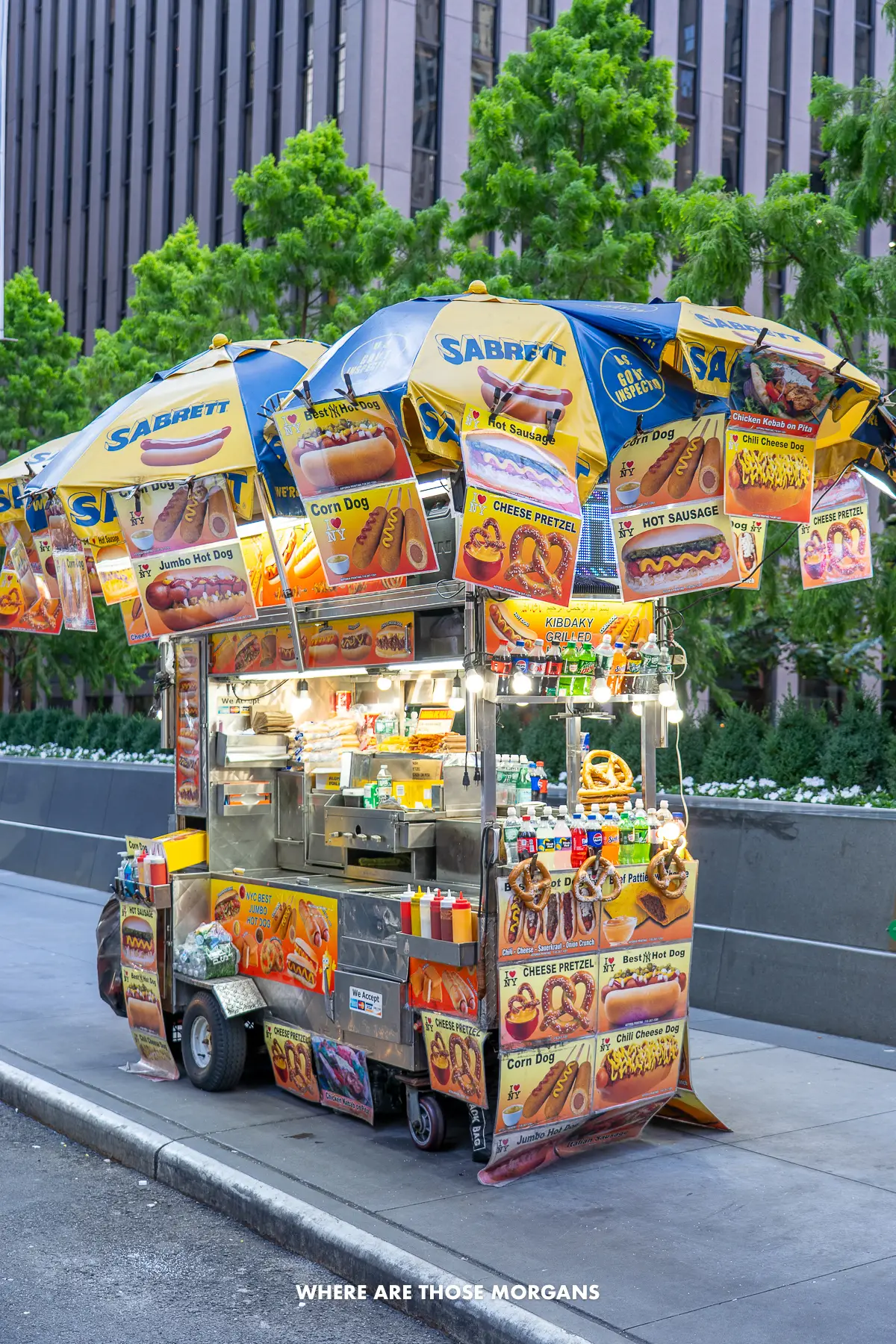 Hot dog stand with umbrellas and lots of snacks outside a building in NYC with green leaves on top of a short wall