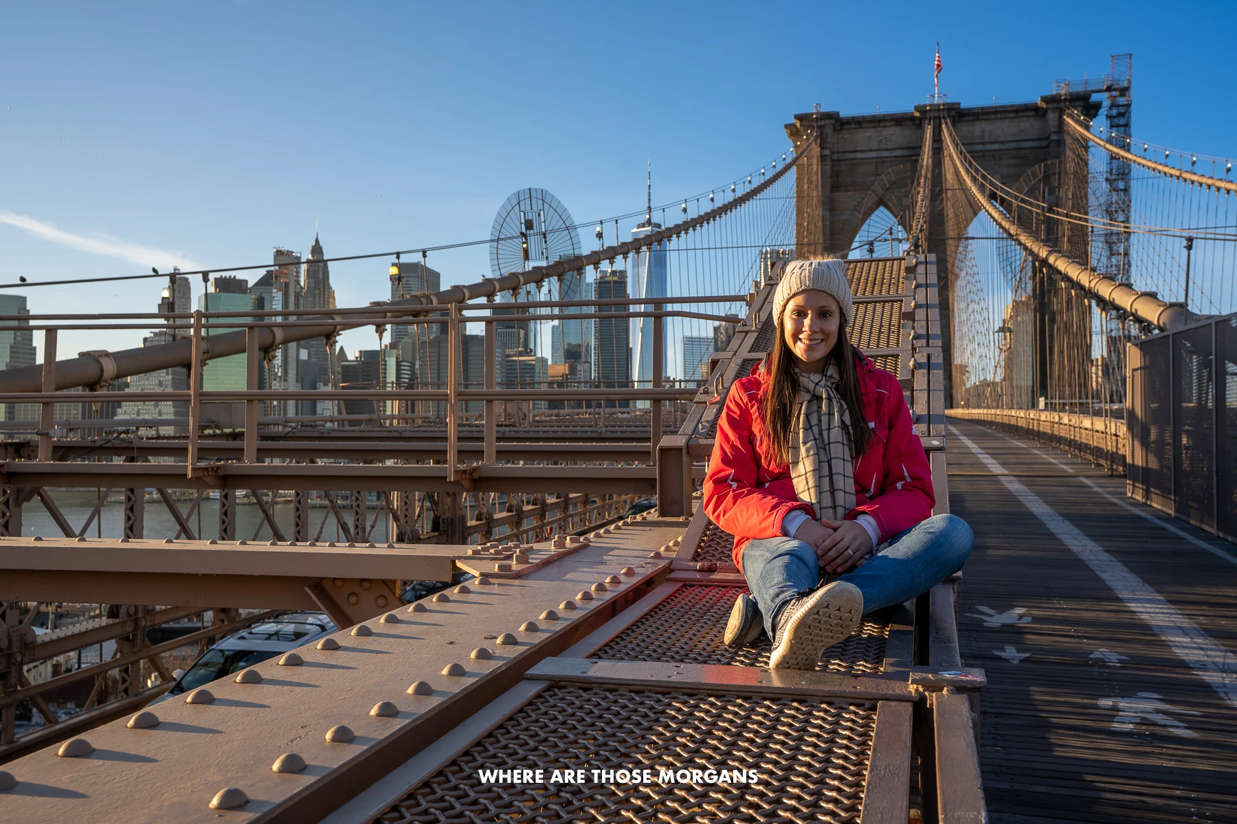 Kristen from Where Are Those Morgans sat on the metal Brooklyn Bridge wearing a coat and hat at sunset