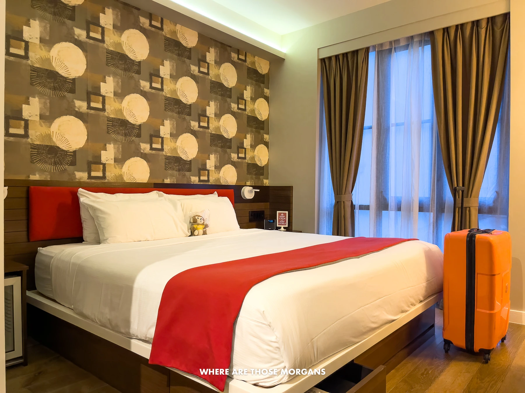 Inside a hotel room called NobleDen in Little Italy NYC with wallpaper behind a bed and a suitcase in front of windows with curtains drawn